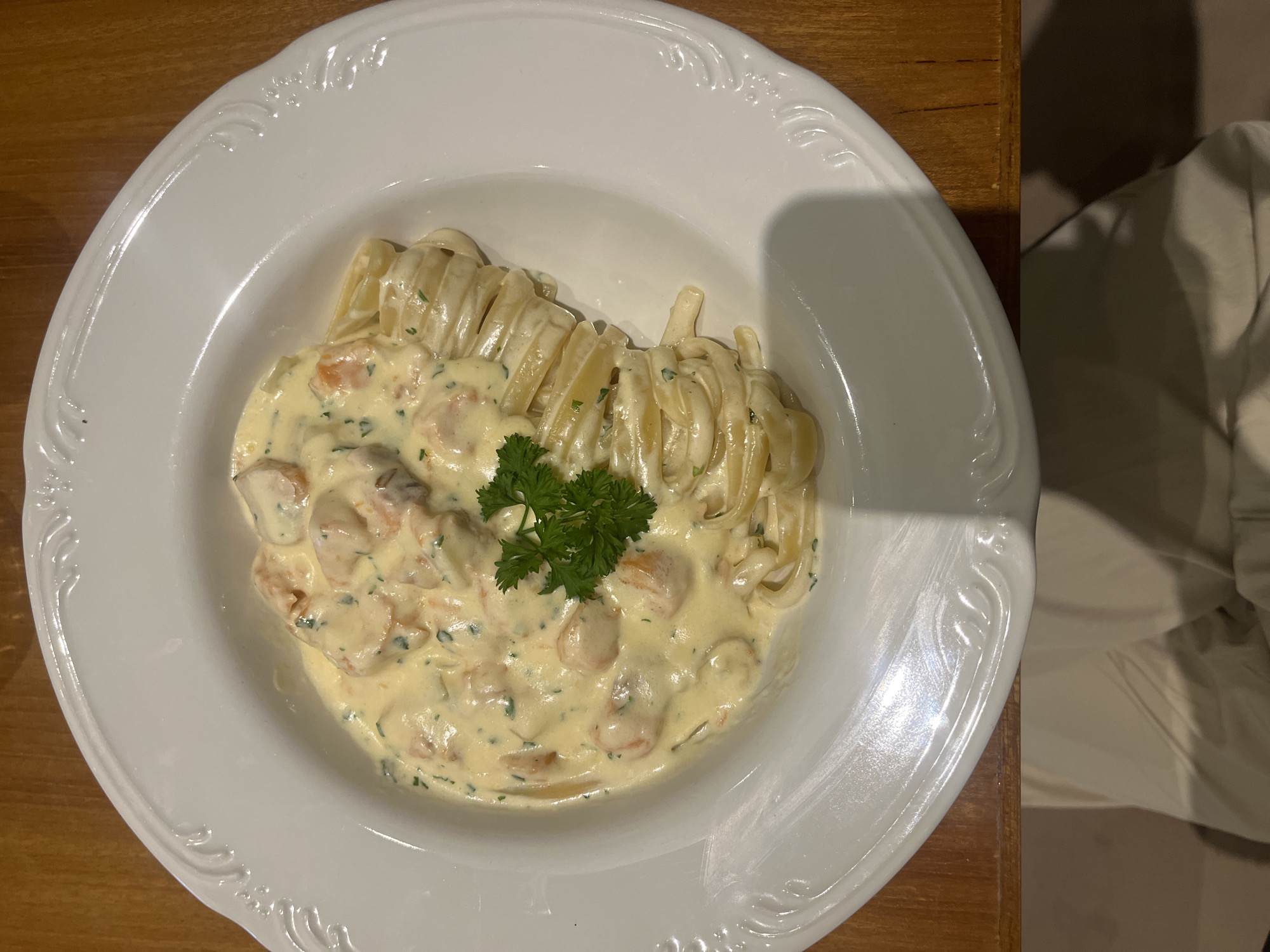 The image displays a close-up, overhead view of a well-presented dish of pasta served in a white, round plate with an ornate, embossed rim. The plate is positioned on a wooden table surface, which shows a distinct grain.
The main subject is the pasta, which appears to be a type of flat, wide noodle, possibly fettuccine or tagliatelle, arranged neatly in the bowl section of the plate. It is generously coated in a creamy, pale yellow sauce that contains visible pieces of seafood, likely shrimp or prawns, and possibly other ingredients like mushrooms or scallops. The sauce also has flecks of green herbs, suggesting parsley or chives. A fresh sprig of bright green parsley garnishes the center of the dish, adding a pop of color.
The lighting appears to be artificial, casting a soft shadow from the rim of the plate on the background to the right. To the right of the plate, a blurry, light-colored object, possibly a chair or draped fabric, is visible in the background. The scene suggests an indoor setting, likely a restaurant or a dining area at home. There is no human activity depicted, nor is any text visible in the image.