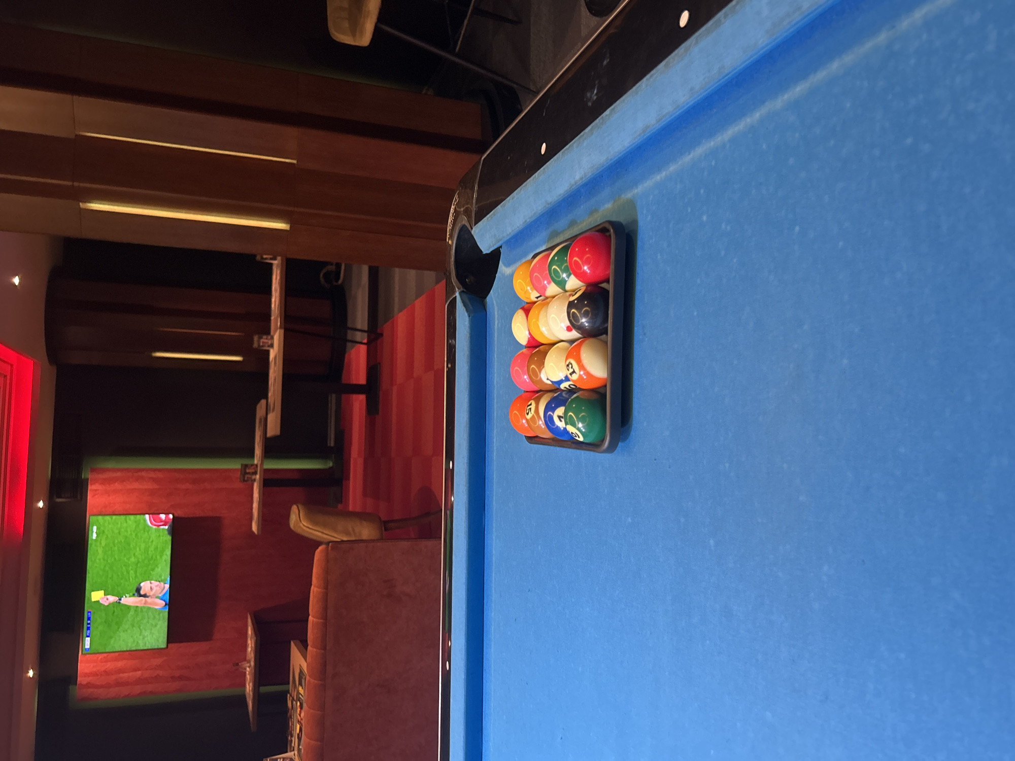 The image shows a partial view of a blue pool table. A rack containing pool balls sits near the edge of the table. The background reveals a room with a television displaying a soccer match. The setting appears to be a bar or entertainment venue, possibly in Sofia, Bulgaria. Furniture, red patterned walls, and interior lighting details are visible in the background.