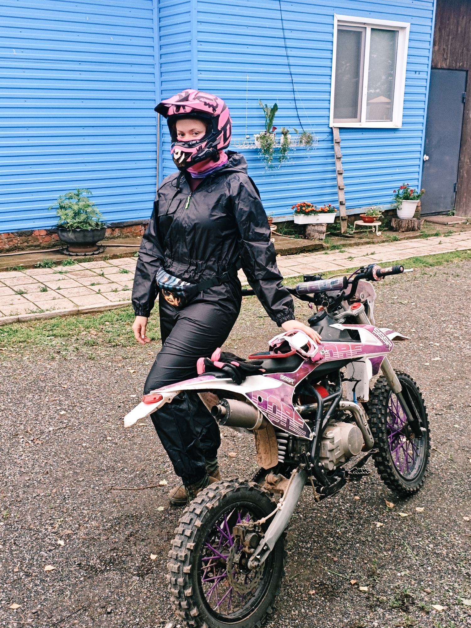 In front of a bright blue corrugated metal building in Rassol'naya, Russia, a person stands beside a dirt bike. The person, wearing a pink motocross helmet, black jacket, and black pants, has a fanny pack around their waist. The dirt bike is white and pink with purple rims. The ground is covered in gravel and scattered leaves. The building has a white-framed window and a dark gray door. Several potted plants are visible around the building.