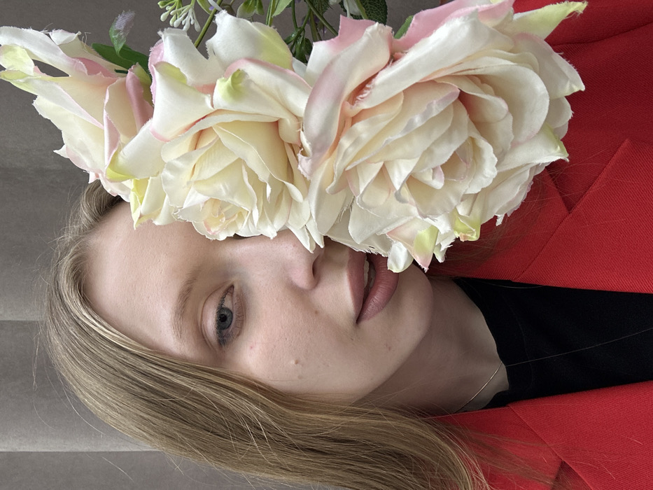 A close-up portrait depicts a young adult female situated in Chaikovsky, Russia. She possesses long, straight blonde hair and blue eyes, with a light complexion exhibiting minor skin variations. She wears a red blazer over a black top, complemented by a thin silver-colored necklace. The left half of her face, including her left eye, nose, and mouth with slightly parted lips, is visible and directed towards the viewer. The right side of her face is deliberately obscured by a prominent arrangement of several large, artificial off-white roses with pink-tinged edges and faint green accents, accompanied by smaller green foliage and tiny white floral elements. The background consists of a uniform, light grey curtain or fabric.