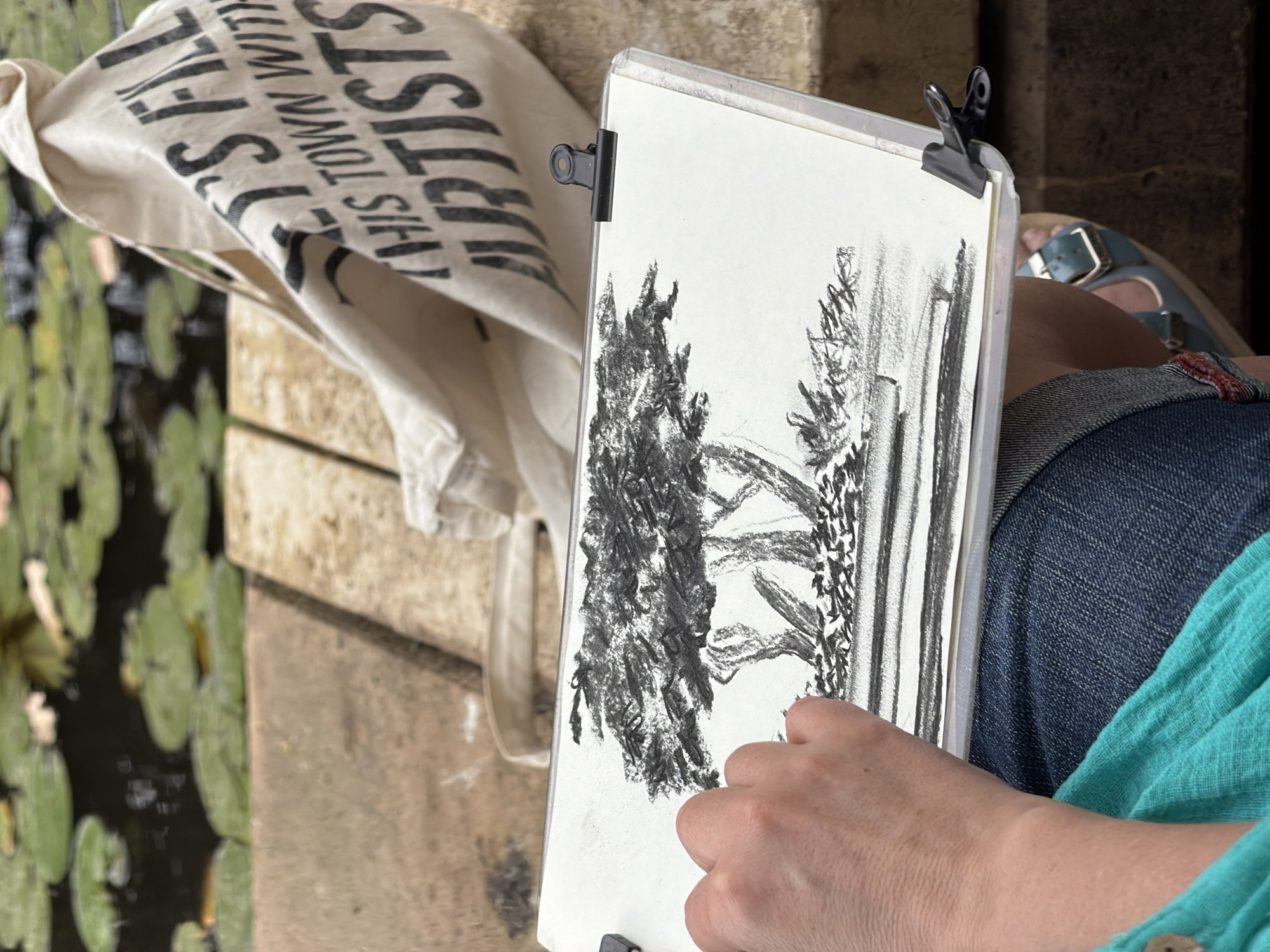 A person is sketching in a park, likely in Tel Aviv, Israel. The drawing, done in charcoal on paper, depicts a landscape with trees. The artist is wearing jeans and a turquoise shirt and sits with the sketchbook resting on their lap. The person is right-handed. A canvas bag with the words "This Town With Artists" hangs nearby. In the background, a body of water is visible, covered with lily pads. The setting appears to be a park or garden, with the person sitting on a wooden structure near the water.