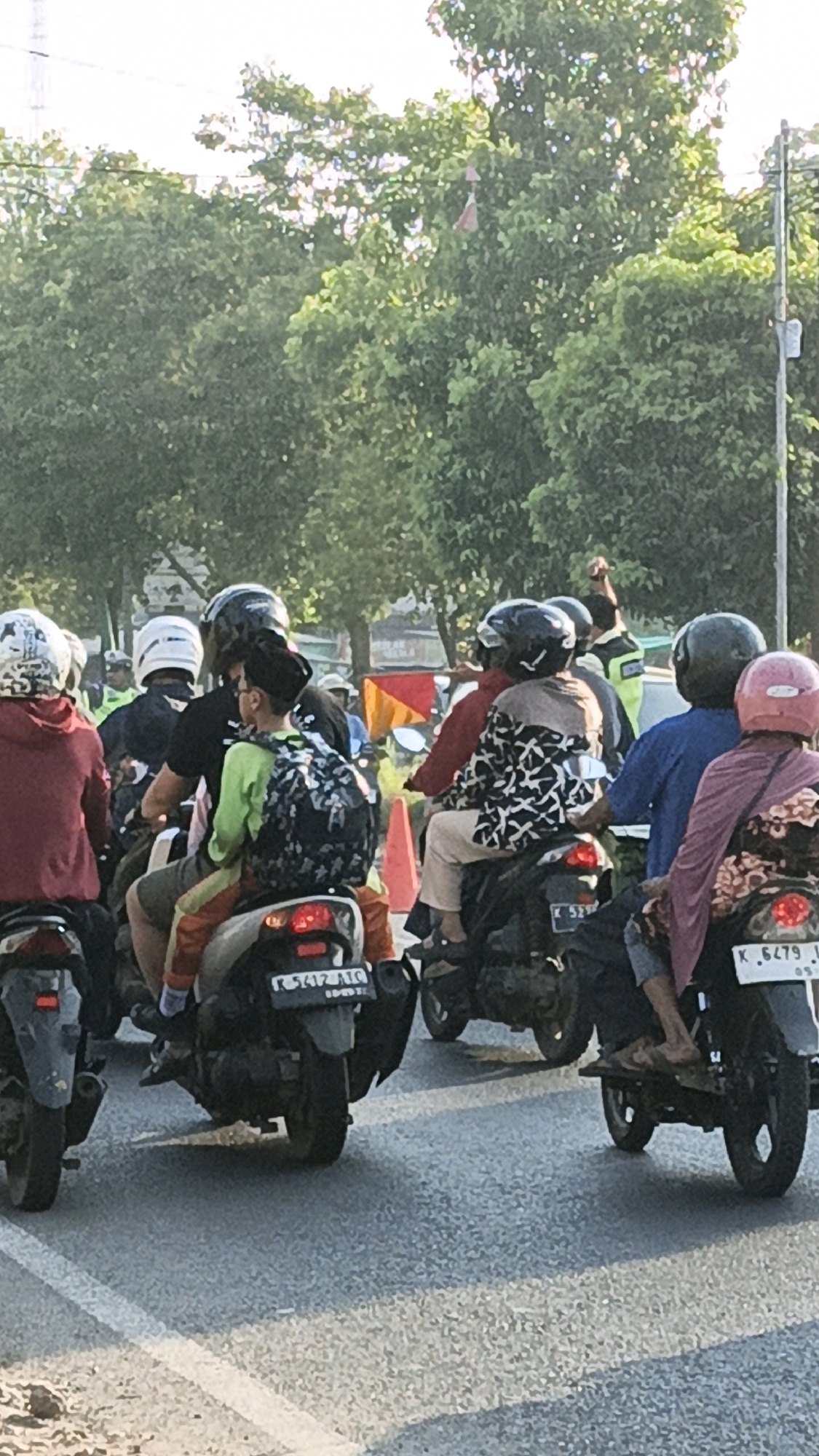 The image captures a scene of several motorcycles on a paved road, likely in Kuwasen, Indonesia. The motorcycles are carrying multiple passengers, with most individuals wearing helmets for safety. The people are dressed in casual clothing. In the background, lush green trees line the road, and a power pole is visible on the right side. The road surface appears to be asphalt, with white lane markings. The lighting suggests it is daytime, possibly morning or late afternoon, with sunlight casting shadows. The overall impression is a typical mode of transportation in the region, with a focus on practicality and community travel.