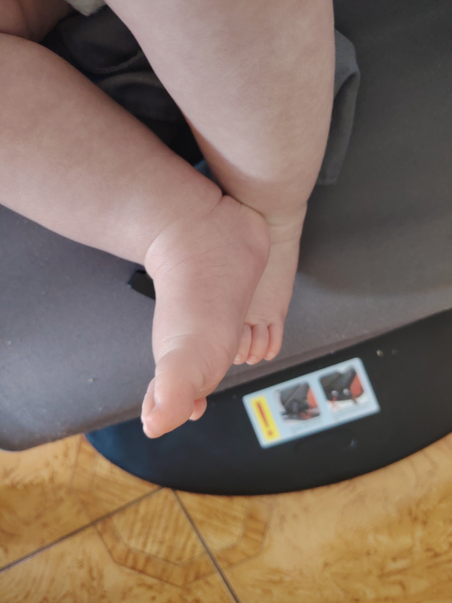 A close-up, high-angle shot shows the legs and feet of a baby. The baby's skin is pale and smooth, with visible creases on the ankle and toes. The baby is wearing a grey garment that partially covers their legs. The baby is positioned on a dark, flat surface, possibly a car seat or a mat, which is resting on a wooden floor. The floor has a parquet pattern with light brown and darker brown tiles. In the lower right corner, a small, rectangular sticker is visible on the dark surface, featuring two illustrated images of a car seat with red accents and a yellow exclamation mark. The image is likely taken indoors. There is no information to determine the location in Konakovo, Russia.