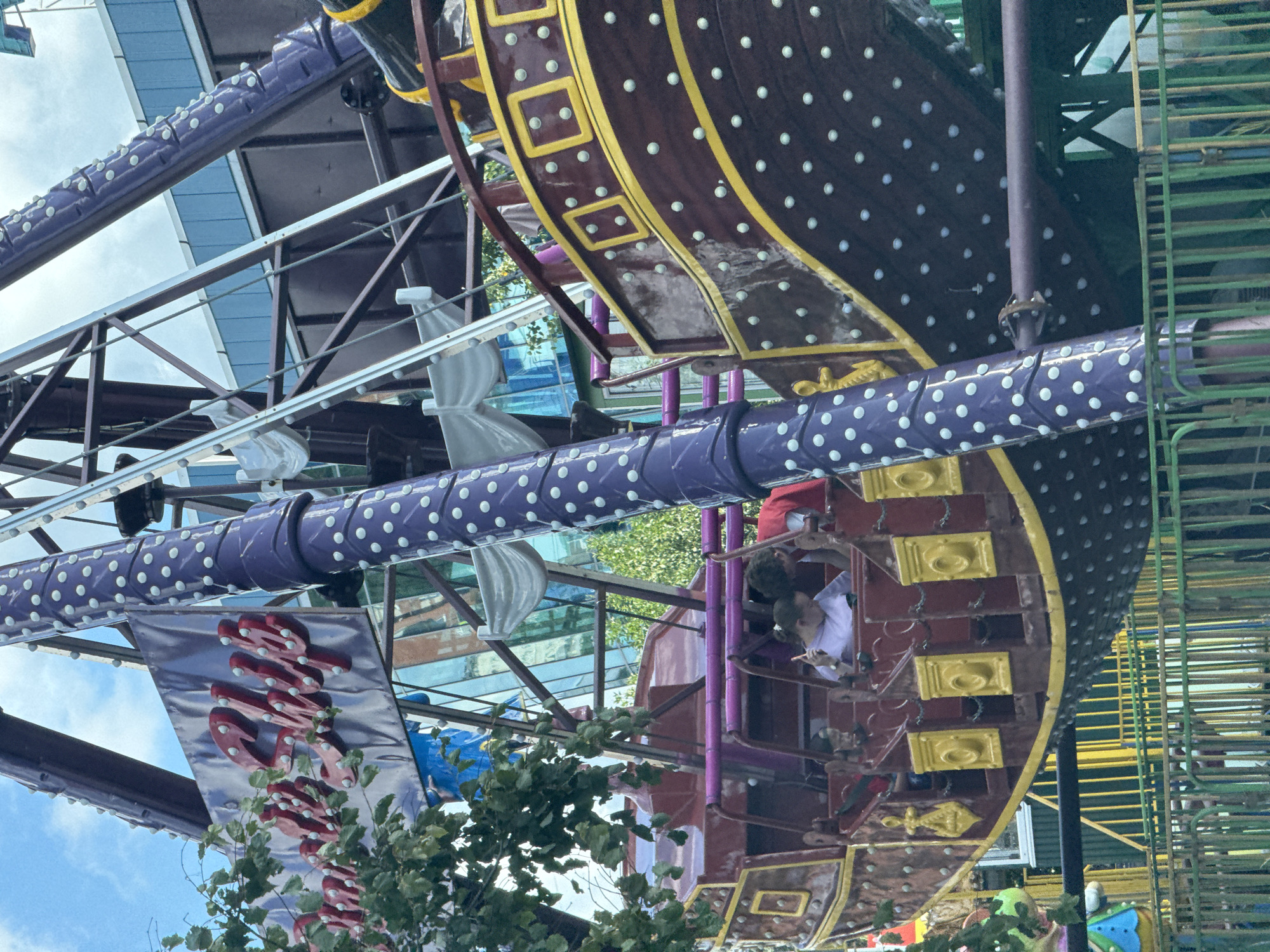The image shows a close-up of a carousel attraction designed as a pirate ship, likely located in an amusement park. The ship is primarily maroon with gold trim and yellow decorative elements, featuring numerous small, white, circular lights along its hull and supporting structure. A large, purple, polka-dotted pole is prominently visible on the left, part of the ride's mechanism. Two individuals, a young woman in a light purple top and a young man in a red shirt, are seated in the ship's passenger area, with the woman holding a smartphone. Behind the ship, elements of a building with a blue facade and reflective windows are visible, along with trees and a cloudy sky. The foreground shows a green metal fence. The context of Sinyavino, Russia, suggests this amusement park might be a local attraction within the town.