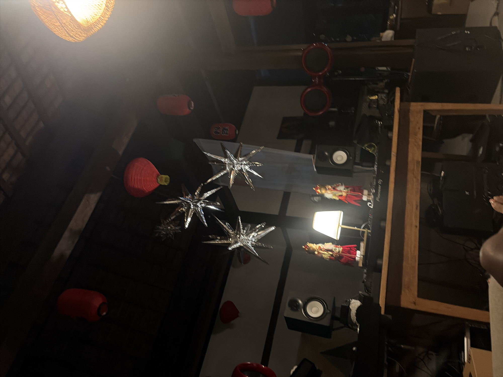 The image depicts an indoor scene that appears to be a dimly lit room, possibly a living area or entertainment space. Several red lanterns are hung from the ceiling, along with three silver star-shaped ornaments. In the foreground, a wooden table is set up with DJ equipment, including speakers and a Pioneer DJ controller. Two figures, possibly dolls or mannequins dressed in red and gold, stand on the table. A lamp with a white shade is also present. In the background, a large wicker-like light fixture hangs from the ceiling, casting a warm glow. The overall atmosphere suggests a festive or celebratory occasion. The location is identified as Kediri, Indonesia, though no specific landmarks or contextual clues within the image directly confirm this.