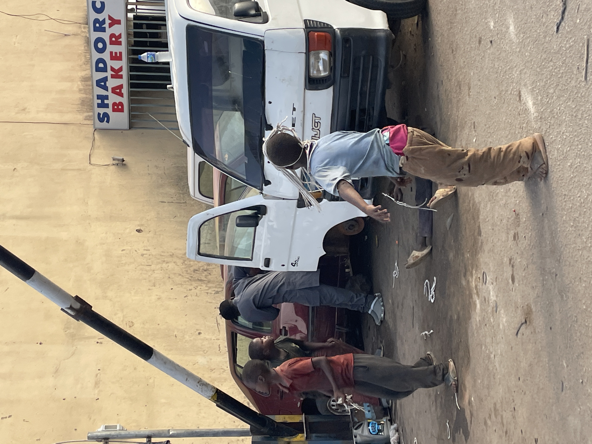 The image depicts a street scene in Jos, Nigeria, with several individuals gathered around vehicles, seemingly engaged in repair work. A white van and a maroon car are the primary vehicles in view, with at least three individuals present. One person, a child, is standing near the white van with a makeshift headpiece made of string. Another individual is bending over the maroon car, while another is standing nearby. The environment appears to be a roadside repair shop, with tools and car parts scattered on the ground. A sign for "SHADORE Bakery" is visible on a nearby building. A pole with black and white stripes is in the foreground.