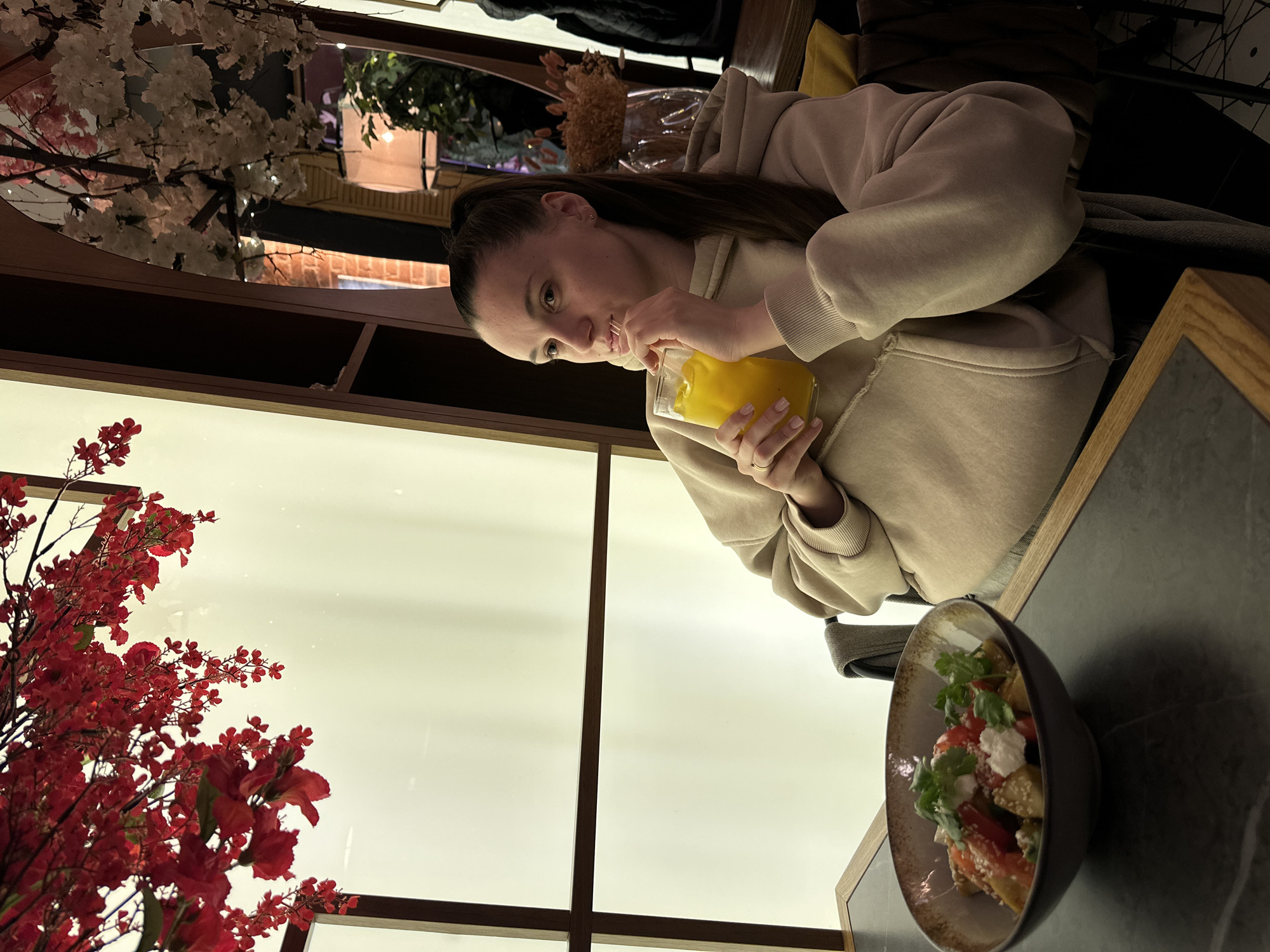 A woman is seated at a table indoors, drinking an orange beverage with a straw. On the table is a bowl of salad. The environment includes floral decorations and a window with a view to the outside. The location is likely a restaurant. The city of Ovoshchnoy, Russia is not verifiable from this image.
