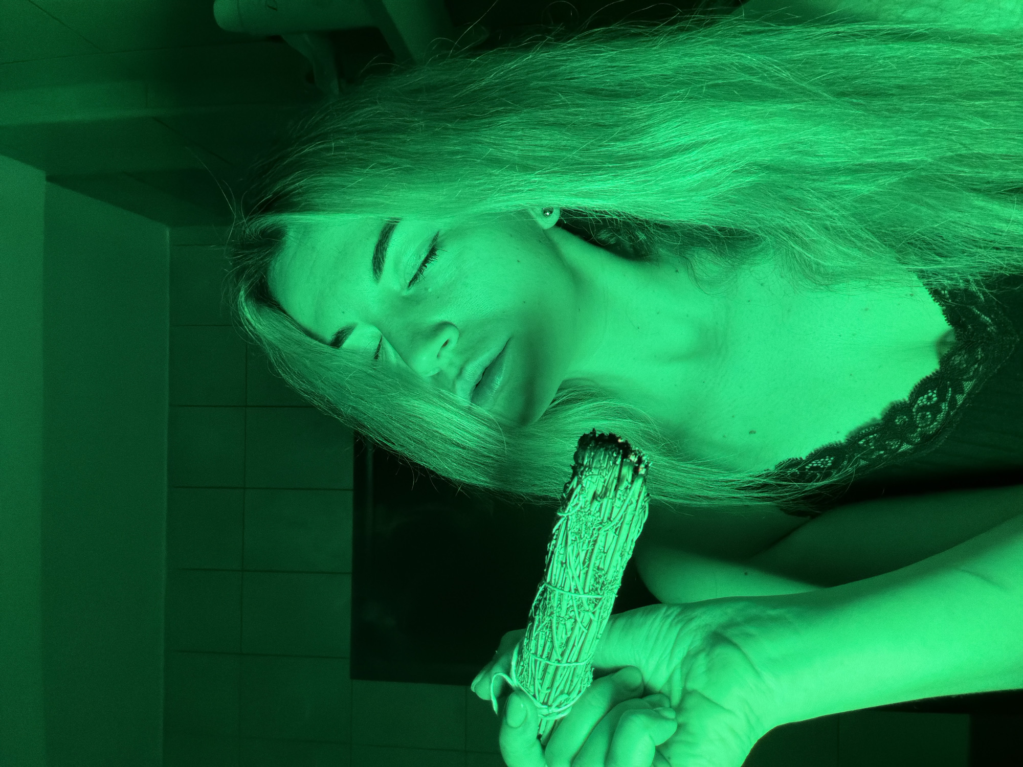 A woman with long blonde hair, eyes closed, holds a smoking bundle of sage in her right hand. She is positioned indoors with a tiled wall visible. The image has a strong green color cast. No location is evident.