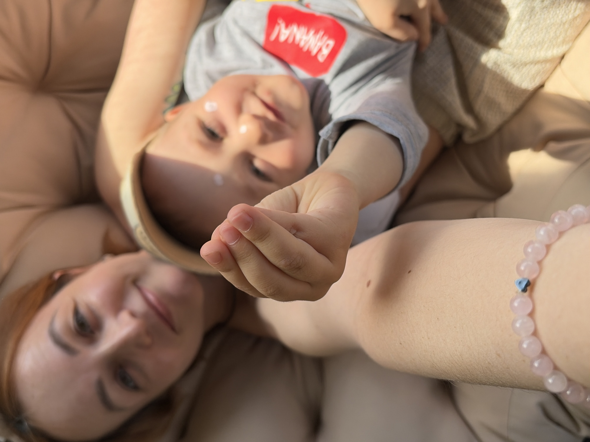 The image shows a selfie taken by a woman with a child in Chaikovsky, Russia. The woman is lying down on a beige cushion or padded surface, with the child positioned on top of her. The child is wearing a gray shirt with the word "BANANA!" printed on it. Both the woman and the child have fair skin. The child appears to have sunscreen or a similar cream applied to their face. The woman is wearing a pink beaded bracelet with a blue heart charm. The overall lighting suggests the photo was taken outdoors.