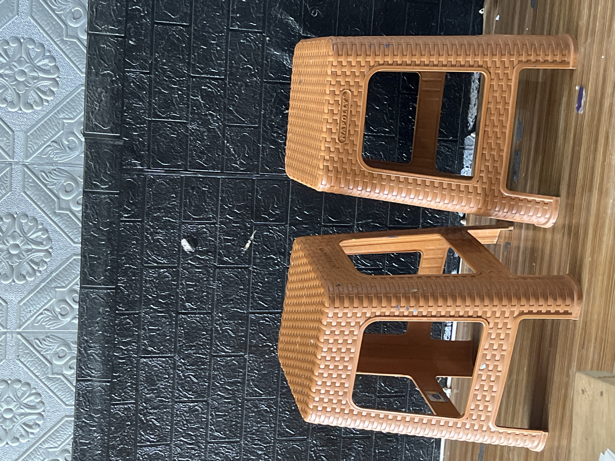 Two orange plastic stools are placed side-by-side in front of a black brick-patterned wall. The wall has a white textured pattern on top. The stools have a woven texture and are square with cutouts on the sides. One stool has the word "NAPOLLY" imprinted on its seat. The floor is made of wood planks. The scene appears to be indoors, possibly in a shop or an outdoor seating area in Sejahtera, Indonesia.