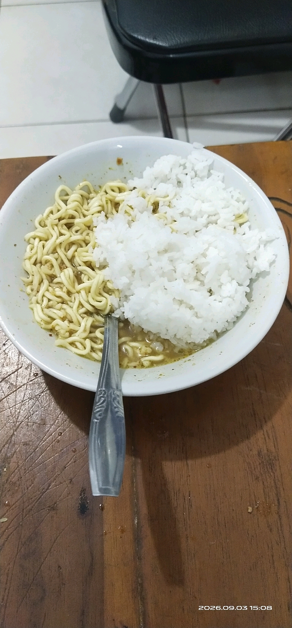 A top-down, close-up shot shows a white bowl filled with instant noodles and white rice. The bowl is placed on a wooden table with a visible grain pattern. A metal spoon with an ornate handle rests in the bowl, partly submerged in the noodles and broth. The background is slightly out of focus, revealing a black chair and a tiled floor. A timestamp reading "2026.09.03 15:08" is visible in the bottom right corner of the image. This scene is consistent with a meal being served or consumed in a domestic setting in Tamiang Layang, Indonesia.