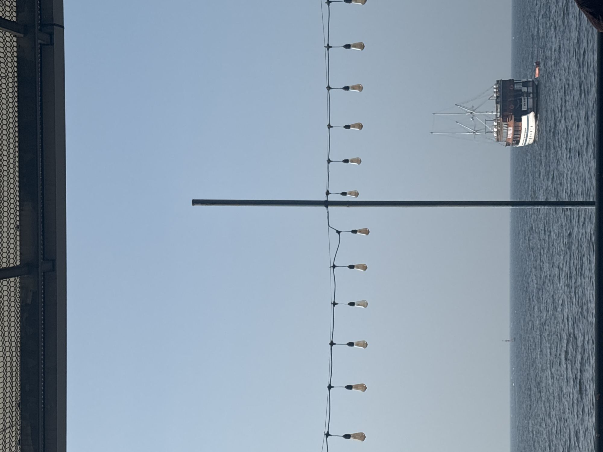 This image captures a view of the sea in North Jakarta, Indonesia, possibly from a waterfront establishment. In the foreground, a series of Edison-style light bulbs hang suspended from a wire, strung horizontally across the frame. A tall, slender pole stands vertically near the center, extending upwards from below the frame. The ocean stretches out in the midground, with gentle waves visible. On the water, a multi-decked wooden boat with white trim and masts is anchored. A small orange inflatable boat is attached to its stern. In the far distance, a solitary buoy marks the horizon. The sky above is a clear, pale blue. A textured metal structure is visible at the very top of the image, suggesting the presence of a roof or awning.