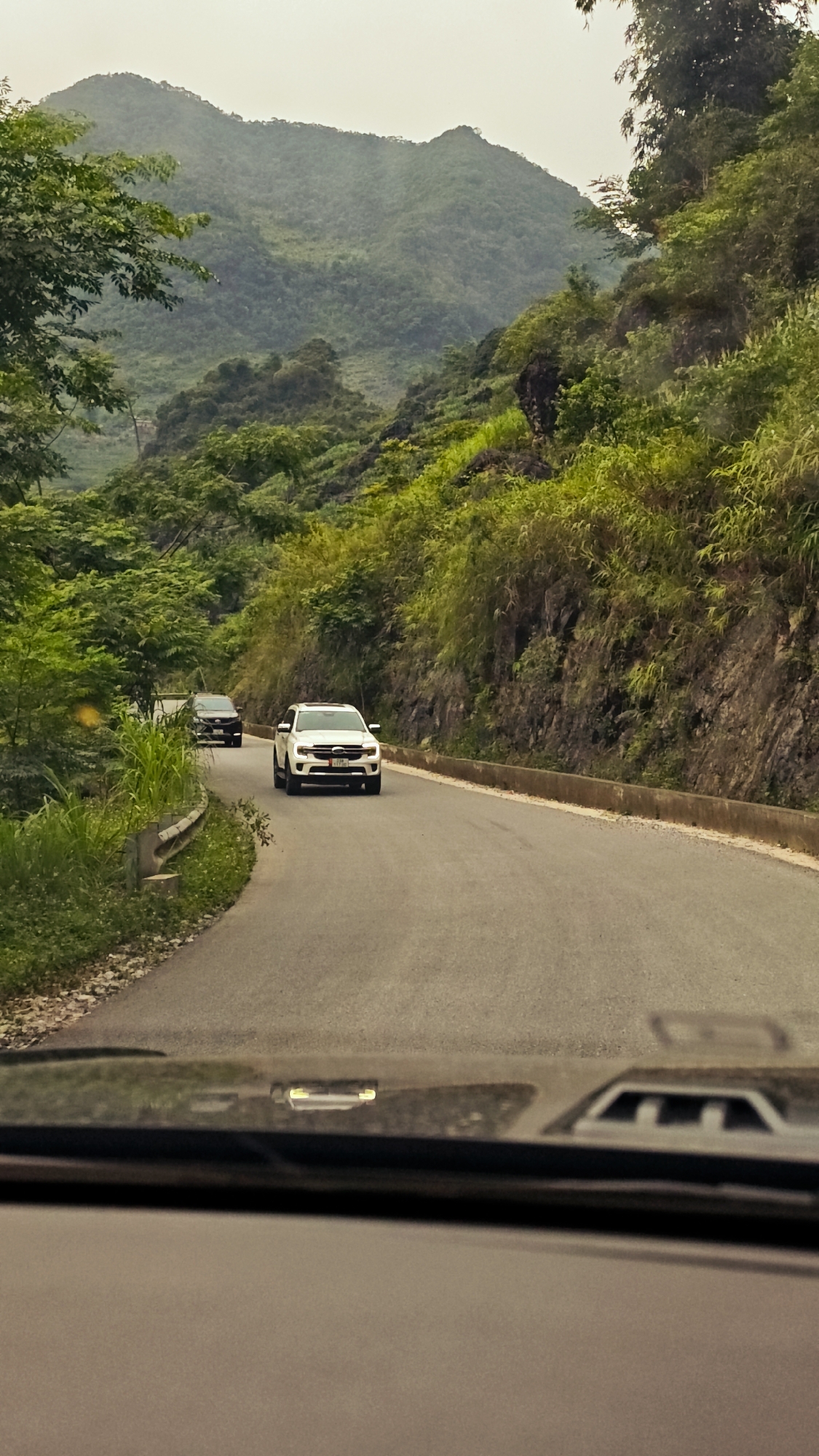 The image depicts a winding road in the Cán Tỷ Commune, Vietnam, as viewed from inside a moving vehicle. Two cars are visible ahead on the road: a white SUV in the immediate foreground and a dark-colored car further behind. Lush, green vegetation covers the surrounding hills and mountains. The road is asphalt and has concrete barriers on one side. The sky is overcast.