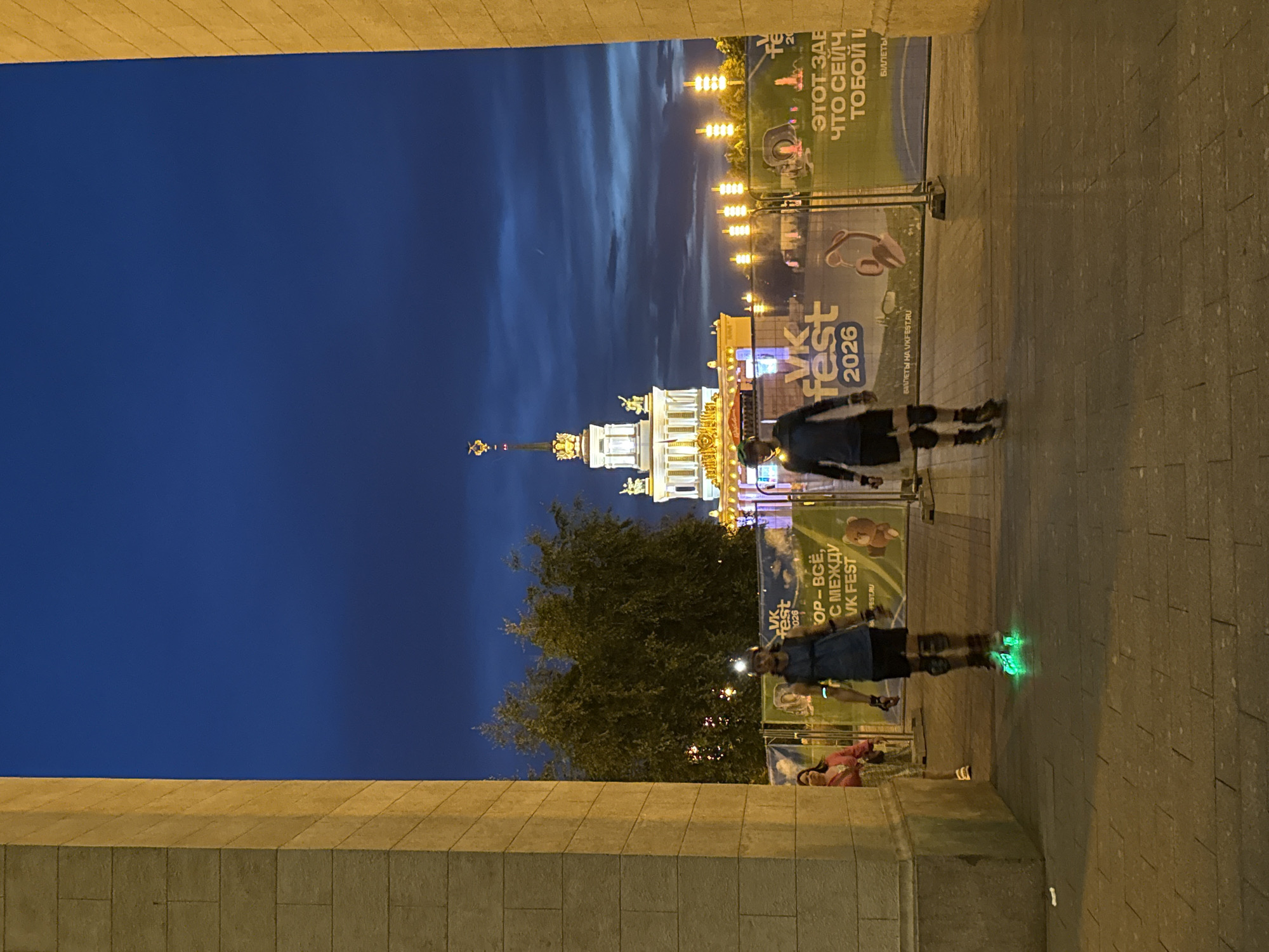 This image depicts an outdoor scene at night, framed by two large, light-colored stone archways. In the background, a prominent, ornate building with a spire, reminiscent of Soviet-era architecture, is illuminated. A dark blue sky with some clouds stretches above. In the mid-ground, two individuals are rollerblading. The person on the left is wearing a dark outfit and a helmet with a light. The person on the right is also wearing dark athletic clothing, knee pads, and rollerblades with bright green illuminated wheels. They are both facing away from the viewer and moving towards the illuminated building. Several banners with Russian text and graphics are visible behind the rollerbladers, indicating an event or festival, possibly "VK Fest 2026," as suggested by the text on some banners. To the left, a woman in a pink top and patterned skirt is partially visible, standing still. The ground is paved with large, rectangular stones. The scene appears to be set within a public space, possibly an exhibition center or park, given the architecture and the presence of banners and lighting. The building in the background strongly suggests this location is VDNKh (Exhibition of Achievements of National Economy) in Moscow, Russia, specifically the main pavilion, Pavilion No. 1 "Central."
