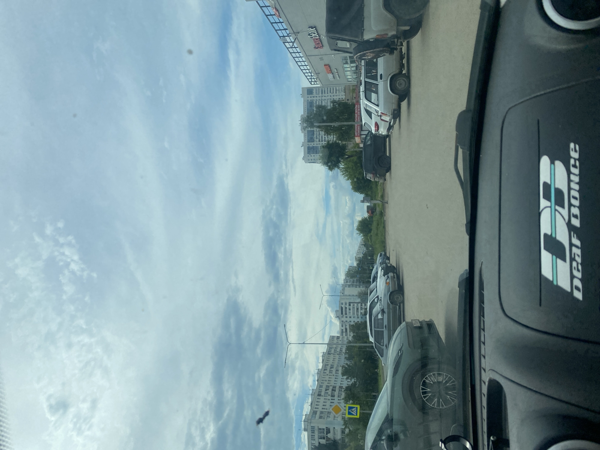 The image, taken from inside a vehicle, shows a street scene with parked cars and buildings. In the foreground, the dashboard of the car is visible, with a logo that says "DEAF BONCE". Outside the car, a row of parked cars is visible, including a dark SUV, a silver sedan, and other vehicles. Beyond the cars, there are several multi-story residential buildings, some with balconies. The sky is cloudy with patches of blue sky visible. A yellow traffic sign with a blue pedestrian symbol is also visible on the left side of the image. The scene appears to be in a residential area of a city. The specific location is identified as being in Samara, Russia.