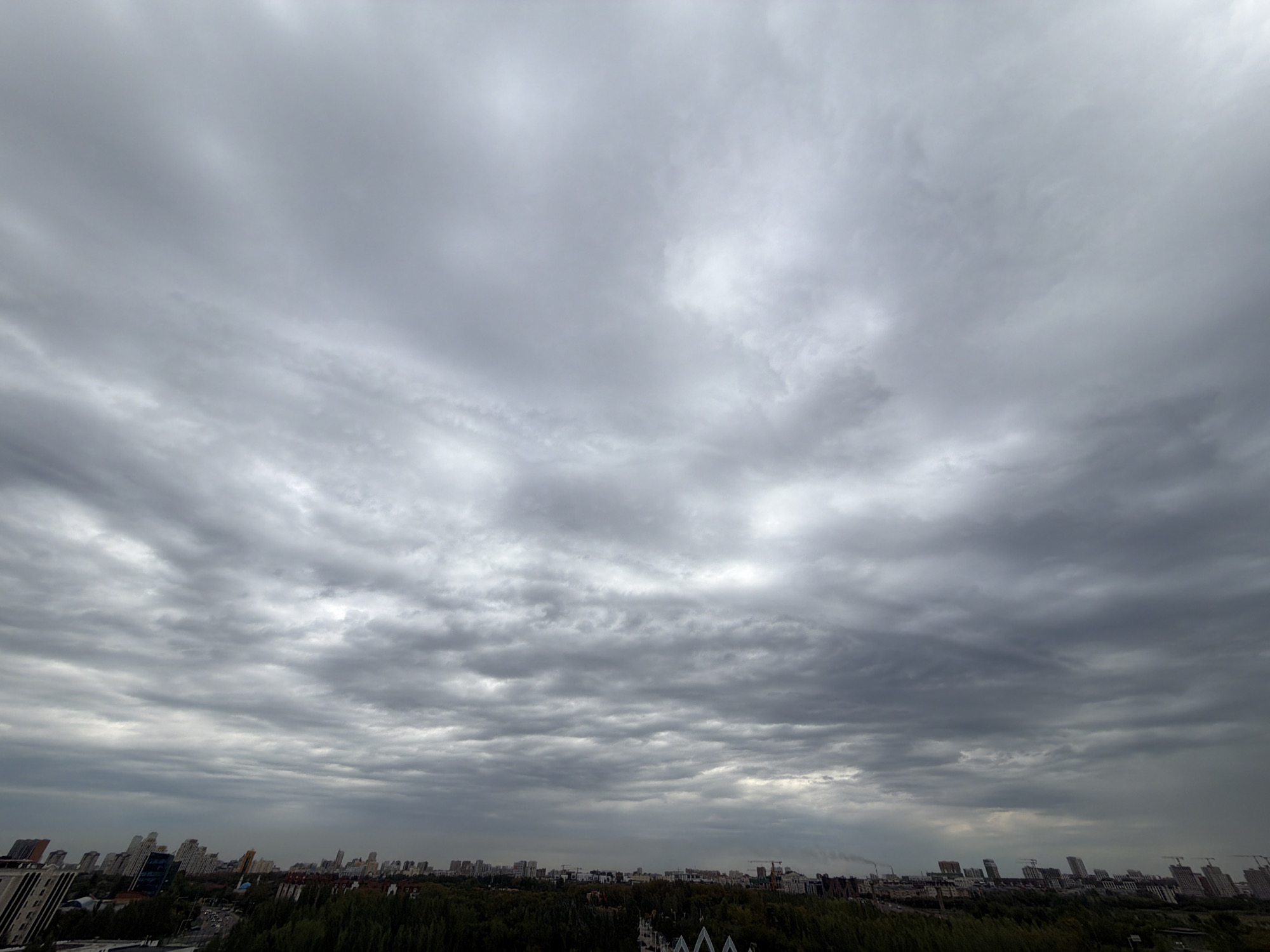 The image depicts a cloudy sky over the city of Astana, Kazakhstan. The sky is filled with dense, gray clouds, suggesting an overcast or potentially rainy day. Below the sky, a cityscape is visible, characterized by numerous buildings, including high-rise apartments and office buildings. Some of these buildings have a modern architectural style, while others appear to be older. A line of trees runs across the foreground, separating the urban structures from the sky. The overall impression is one of a typical urban environment under a somber sky. There are no visible people or specific actions taking place.