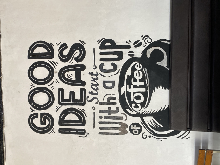 This image depicts an indoor scene in Aluebutuha, Indonesia, focusing on a white wall adorned with a black hand-drawn mural. The mural features the phrase "GOOD IDEAS Start With a cup of coffee" in decorative block and script lettering, accompanied by an illustration of a large coffee cup with the word "coffee" inside. Stylized swirls and small coffee bean motifs accent the design. Below the mural, a dark brown, multi-paneled upholstered bench or booth seating is positioned, obscuring the lower portion of the coffee cup illustration. To the left of the bench, a section of a wooden chair with a silver metal frame is partially visible. The wall surface shows minor imperfections and faint smudges. The upper edge of the wall, where it meets the ceiling, is visible at the top of the frame. No individuals are present in the scene.