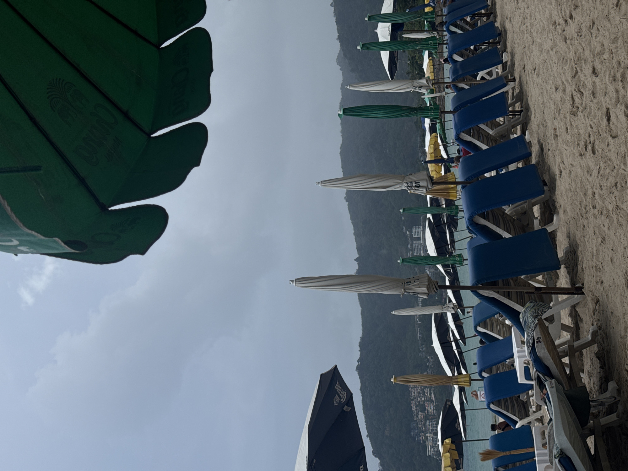 The image depicts a beach in Patong, Thailand, under overcast skies. In the foreground, part of a green umbrella with a company logo is visible, obstructing the view. A row of beach chairs, mostly blue, lines the sand. Umbrellas of various colors including white, green, and yellow, provide shade. In the background, a hillside covered with vegetation is visible, along with buildings. The scene captures a typical beach setting with tourism infrastructure.