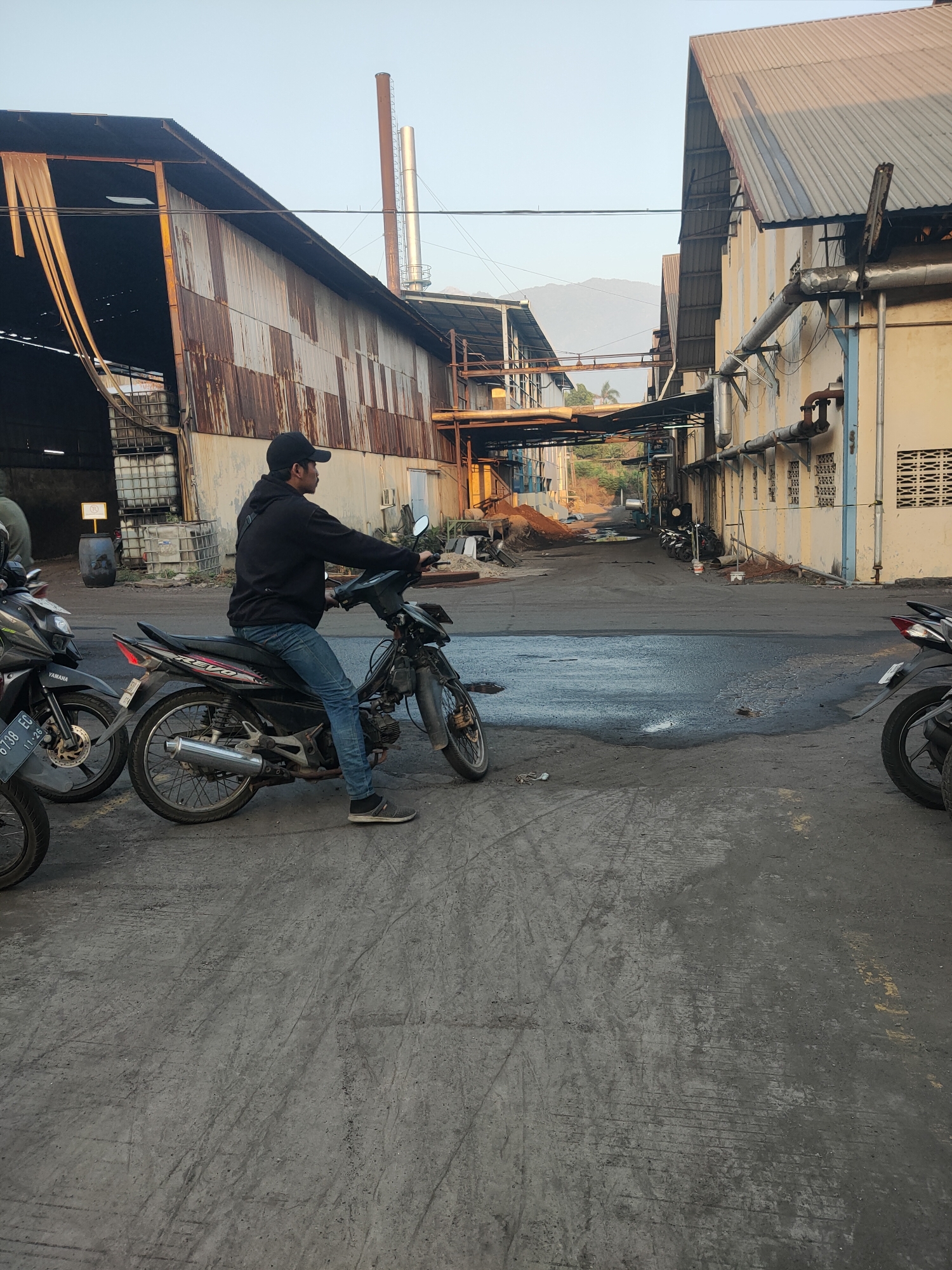 In Beji, Indonesia, a man wearing a black hoodie, black baseball cap, blue jeans, and dark sneakers is seated on a black motorcycle. He is looking to the right. The motorcycle is a "Revo" model, with a silver exhaust pipe and a license plate that reads "6738 EG". The scene takes place in an industrial area with large, weathered metal buildings. One building on the left has a corrugated metal exterior and appears to be open on the side, revealing stacked white plastic containers. Another building on the right is a cream color with yellow accents and features pipes running along its exterior. The ground is paved with asphalt and shows patches of dark liquid and tire marks. In the background, there are more buildings, a tall chimney stack, and distant mountains. Other motorcycles are parked in the vicinity.