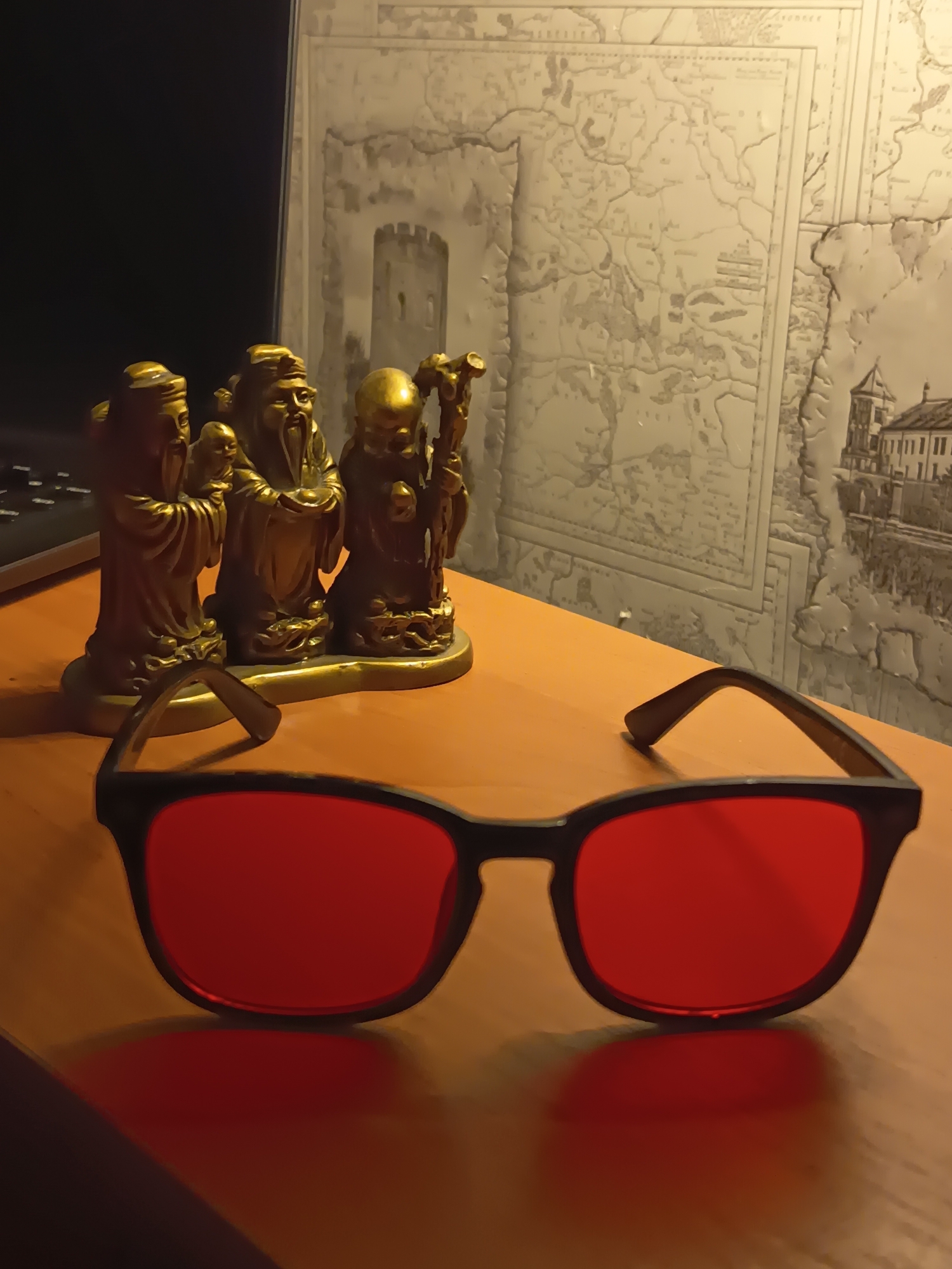 A pair of red-lensed sunglasses rests on a wooden surface in the foreground, with their frames casting a shadow. Behind the sunglasses, three golden figurines of elderly men are arranged on a shelf. The background features a textured wall with a map-like pattern. The scene appears to be indoors, possibly in a home or office setting. The image does not contain any information about the city of Izmail, Ukraine.