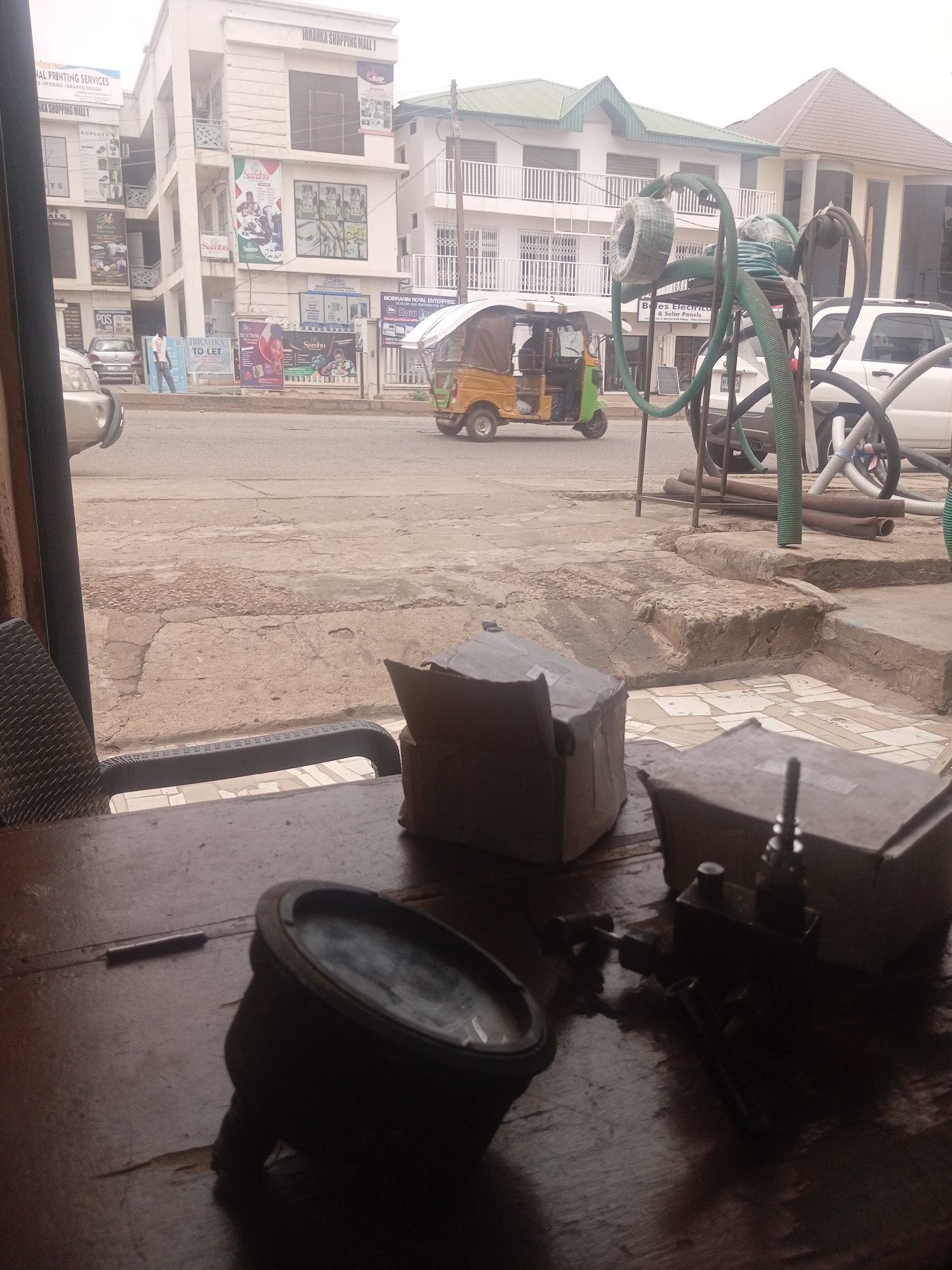 The image depicts a street scene in Sardauna, Nigeria, viewed from an interior location, likely a shop or cafe, looking out through a window. In the foreground, a dark wooden table is visible with several objects, including a round, dark metal object with a clear circular face, possibly a gauge or a lens, positioned on the left. To its right, a small metal piece with a handle and a threaded connection is present, accompanied by a cylindrical metal object and two cardboard boxes, one partially open.
Outside the window, a paved street stretches into the distance. On the left side of the street, several multi-story buildings are present, featuring white facades and balconies. Advertisements and signage are visible on these buildings, including one that reads "IBRANKA SHOPPING MALL 1" and another with "NATIONAL PRINTING SERVICES". A light-colored sedan is parked on the street. A person in a white shirt and blue pants is standing near the sidewalk.
Further down the street, a distinctive three-wheeled vehicle, commonly known as an auto-rickshaw, with a covered passenger area, is in motion. Adjacent to it, to the right, is a white SUV. Behind these vehicles, more buildings with white exteriors and green roofs are visible. In the mid-ground, a collection of green and brown flexible tubes and pipes, some coiled and some lying straight, are stacked on a metal rack or stand. The ground outside appears to be a mix of paved areas, cracked concrete, and some dirt. The overall lighting suggests an overcast or hazy day.