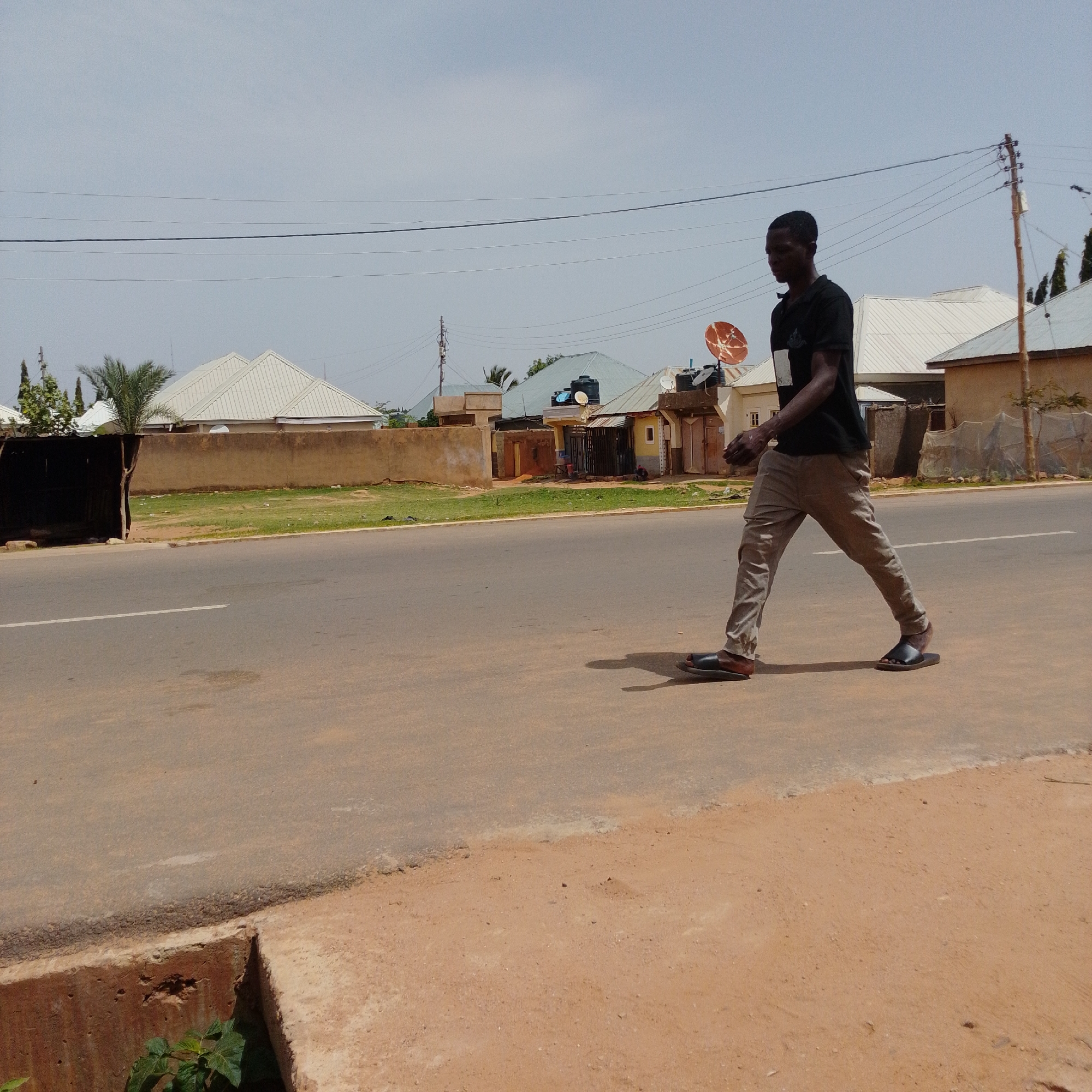 The image depicts a man in a black t-shirt and grey trousers walking on the right side of a paved road in Gombe, Nigeria. He is wearing black sandals. In the background, there are several buildings with corrugated metal roofs, some of which have satellite dishes. A concrete wall separates the road from the buildings, and there is green grass and some vegetation alongside the road. Overhead, power lines are visible against a clear sky. The scene suggests a typical streetscape in a developing urban area.