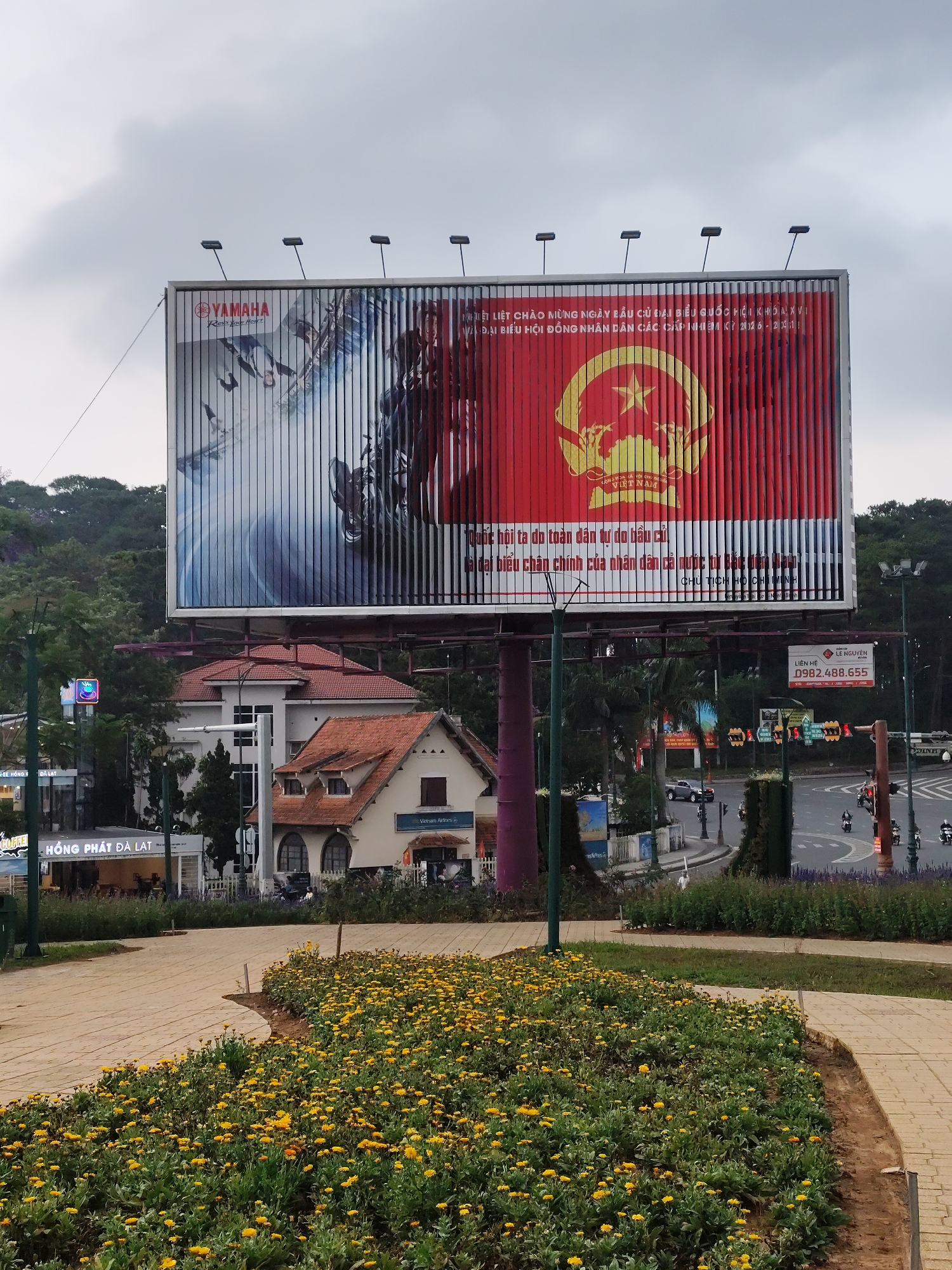 An eye-level shot captures a vibrant urban landscape in Da Lat, Vietnam, under an overcast sky. The central focus is a large billboard mounted on a tall pole. The billboard features vertical louvers, displaying different images depending on the viewing angle.
On the left side of the billboard, an advertisement for "YAMAHA" motorcycles is visible, including the brand's logo and the slogan "Revs Your Heart," along with imagery of a motorcycle. The right side of the billboard displays a significant political message. It features a red background with a large yellow emblem, likely that of the Communist Party of Vietnam, and the text "ĐẢNG CỘNG SẢN VIỆT NAM" (Communist Party of Vietnam). Accompanying text warmly welcomes the election day for the National Assembly delegates (14th term) and People's Council delegates of all levels (2016-2021 term), emphasizing that the National Assembly, freely elected by the people, represents the entire nation, attributed to President Ho Chi Minh.
In the midground, below the billboard, several buildings with various architectural styles are visible, including one with a modern facade and others featuring traditional red-tiled roofs. A busy intersection with moving cars and motorbikes, traffic lights, and streetlights can be seen, indicating an active urban environment. Trees and lush greenery line the roads and surround some buildings. Visible signs include "HỒNG PHÁT ĐÀ LẠT," "COFFE," "Vietnam Airlines," and contact information "LIÊN HỆ 0982.488.655" for "@LE NGUYEN."
The foreground features a paved walkway curving through a garden bed filled with numerous small yellow flowers, adding a touch of natural beauty to the scene. Hills covered in dense foliage form the background. The time appears to be daytime, likely a cloudy or overcast day.