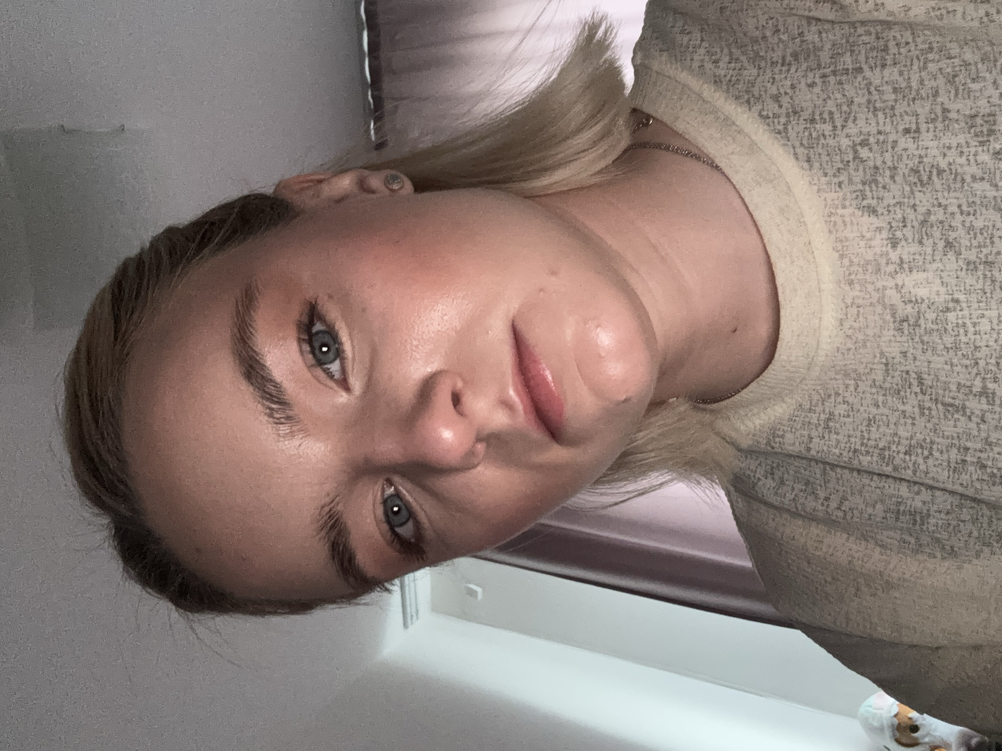 A blonde-haired woman is shown in a close-up headshot, oriented at an angle. She has fair skin, blue eyes, and defined eyebrows. She is wearing a light-colored, textured sweater. A thin chain necklace is visible around her neck. The background consists of a window with closed blinds and a light-colored wall. There is no indication of Vsevolozhsk, Russia in the image. The image shows no actions or interactions.