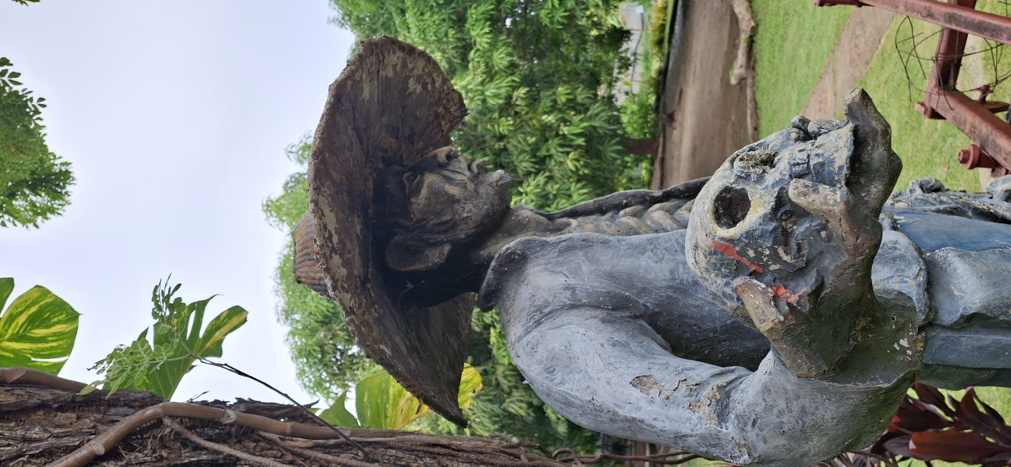A full-body, side-profile, medium shot of a weathered, life-sized statue of a man wearing a wide-brimmed hat stands outdoors in what appears to be a park or garden setting in Vallecito, Venezuela. The statue is depicted in shades of grey, blue, and beige, with visible signs of wear and tear, including chipped paint and texture on its surface. The man, with a stern expression and deeply etched features, is holding a skull in his hands, which are positioned at chest level. The skull is also rendered in a rough, textured material and shows faint red markings. He is wearing a long-sleeved garment that resembles a shirt or jacket, and a woven hat. The background consists of lush green foliage, trees, and a pale sky. A section of a rustic, red metal farm implement is visible in the lower right corner of the frame. The scene is captured in natural, overcast lighting.