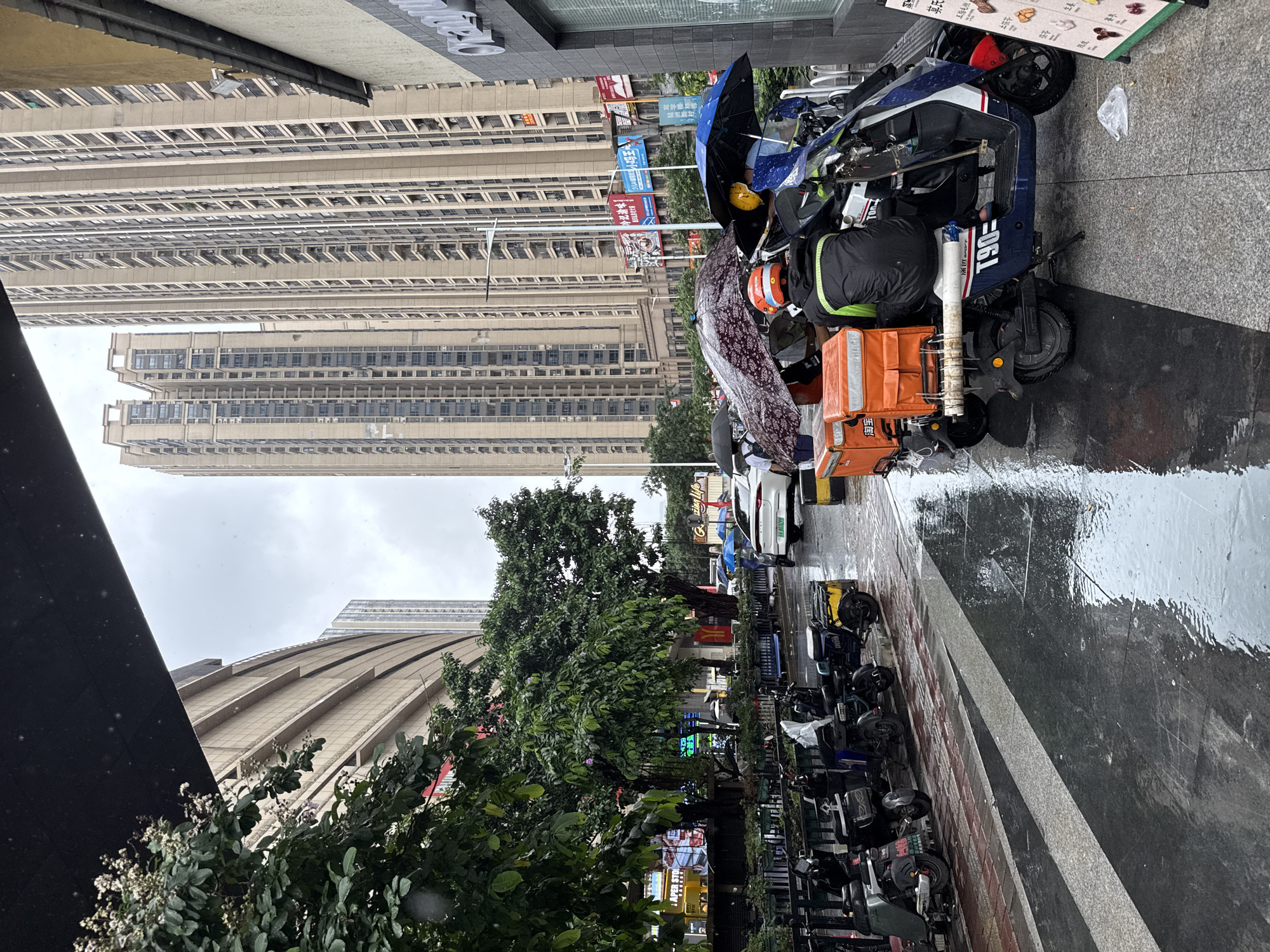 A street scene in Haizhu District, China, shows a rainy day in an urban environment. The foreground features a row of parked electric scooters, some equipped with large orange delivery boxes. One scooter has a rider wearing a black jacket with a green stripe and an orange helmet, sitting on the vehicle under an umbrella. Another scooter is partially visible with a rider under a blue umbrella. The ground is wet and reflective, indicating recent rainfall, with visible streaks of water and scattered raindrops.
Behind the scooters, a sidewalk is lined with trees and a low fence. Beyond that, a street is visible with a white car and a person holding a blue umbrella walking in the distance. The background is dominated by tall, modern apartment buildings with many balconies. The sky is overcast and grey. The scene suggests a commercial or residential area in a densely populated city, with the rain impacting daily activities like deliveries and commuting.