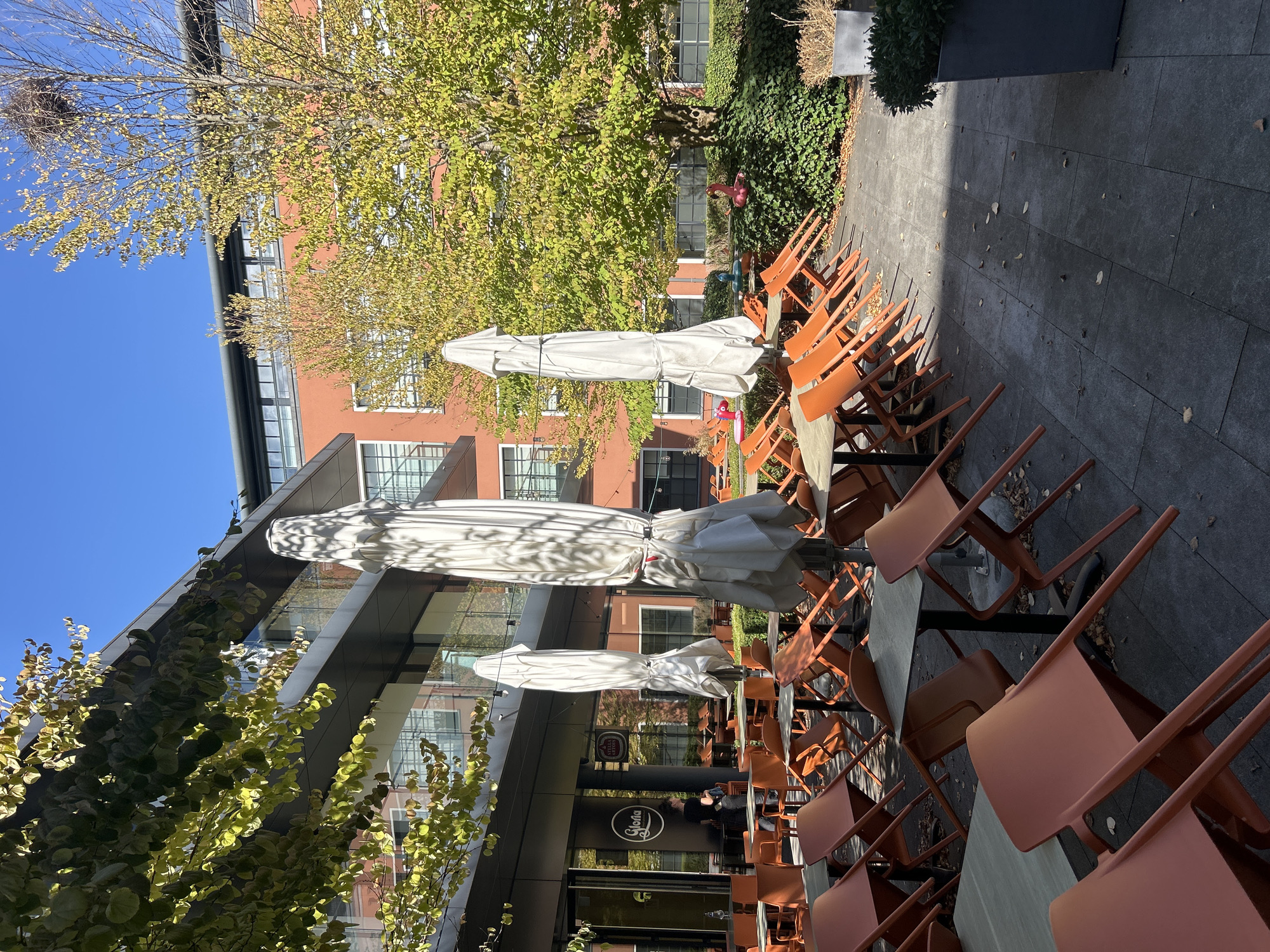 The image shows an outdoor seating area of a restaurant named "Gloria" located in the city of Leuven, Belgium. The scene is bathed in sunlight, casting shadows across the paved ground. Numerous tables and orange chairs are arranged in the outdoor space, with most of the chairs stacked or tipped over, suggesting the restaurant is either closed or preparing for closing. Several large, white umbrellas are closed and standing upright. The building behind the seating area is modern with a glass facade and orange-colored sections. Green trees with yellowing leaves are visible, indicating the season is likely autumn. A person, presumably an employee, is standing near the restaurant entrance, which features large glass doors. In the background, a lush green hedge lines the edge of the seating area, with a modern planter containing dried plants.