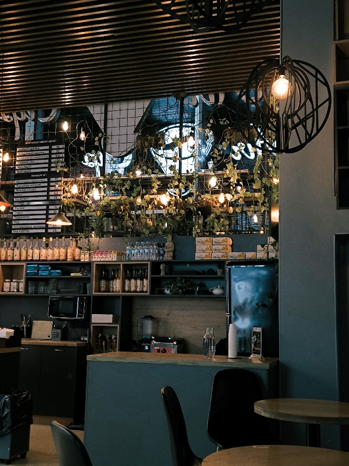 An indoor shot showcases a cafe's interior. A menu board hangs alongside shelves stocked with bottles and various items. Green vines and string lights are draped across a wire mesh background. A refrigerator stands near the counter, while tables and chairs occupy the foreground. A decorative, spherical light fixture is hanging.