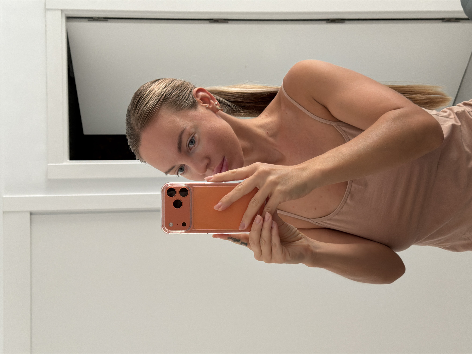 A blonde woman with her hair in a ponytail is taking a selfie in a mirror. She is wearing a peach-colored camisole and holding an orange phone in her hands. She has on small earrings. The background appears to be an indoor setting with white walls and a partially open door. There is no indication of the city of Finestrat, Spain.