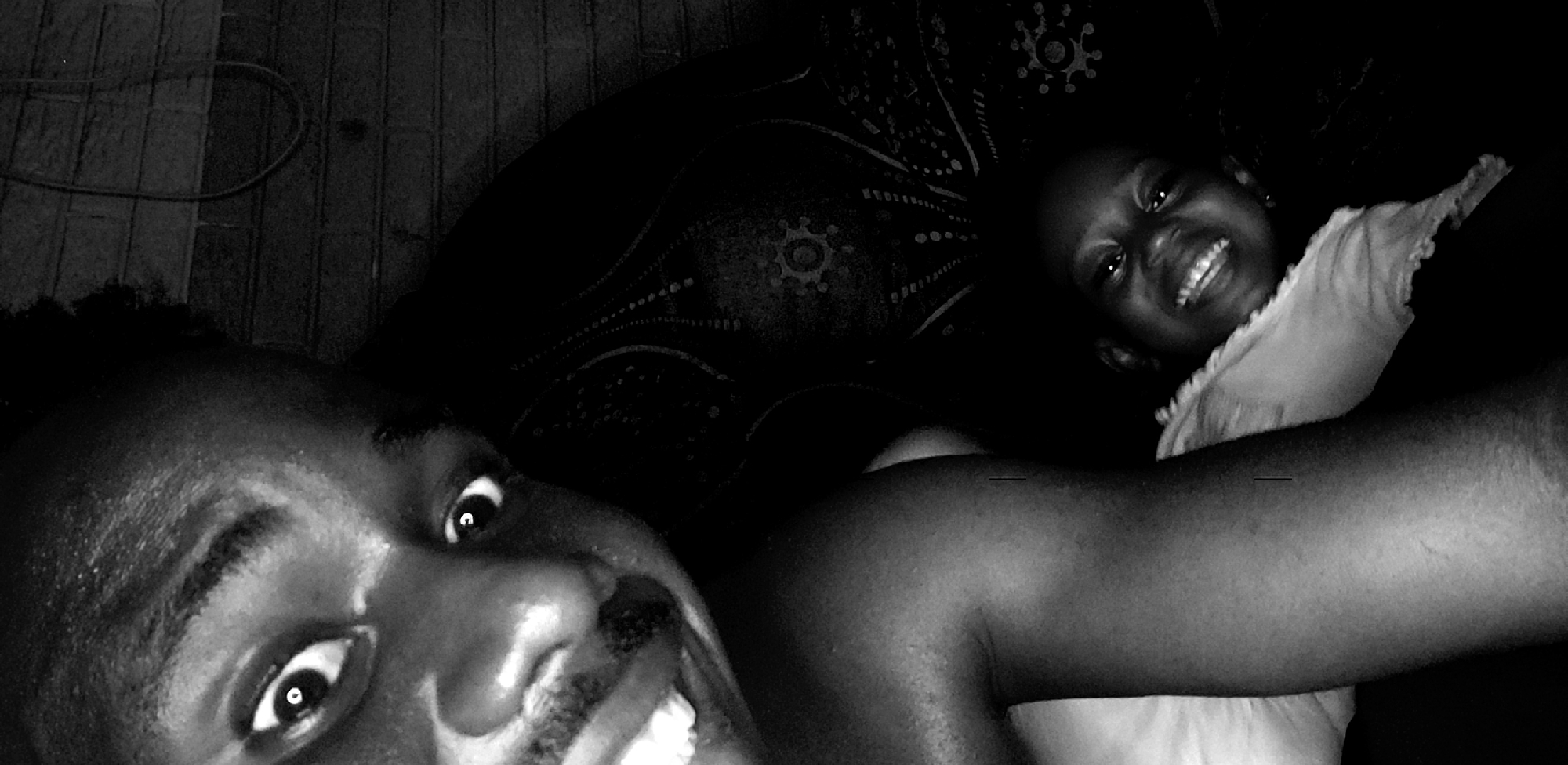 A black and white, close-up photograph captures two individuals, an adult and a child, in an intimate indoor setting in Narayi, Nigeria. The image is taken from a tilted, overhead perspective, possibly a selfie or a photo snapped while lying down.
In the foreground, an adult, likely male given the hint of facial hair, occupies the lower left and central parts of the frame. Their face is turned towards the left, with wide, bright eyes looking directly at the camera. A broad, joyful smile reveals their teeth. A dark, textured surface, possibly hair or a dark fabric, is visible above their forehead. The top left corner shows dark wooden floorboards or wall panels with a thin cable.
Towards the upper right of the frame, emerging from the shadows, is the face of a child, likely female. The child also looks towards the camera with a wide, bright smile, conveying happiness. They are wearing a light-colored garment, possibly a t-shirt or nightgown, with ruffled details around the neckline. A dark, patterned fabric, resembling a blanket or bedspread with circular and dotted motifs, is visible around the child and in the mid-ground.
The lighting is very dim, creating strong contrasts between the illuminated faces and the deep shadows, suggesting a nighttime setting or a very dark room. Both subjects display clear emotions of happiness and warmth. An adult's arm is visible extending towards the right side of the frame, possibly embracing the child. No visible text is present in the image.