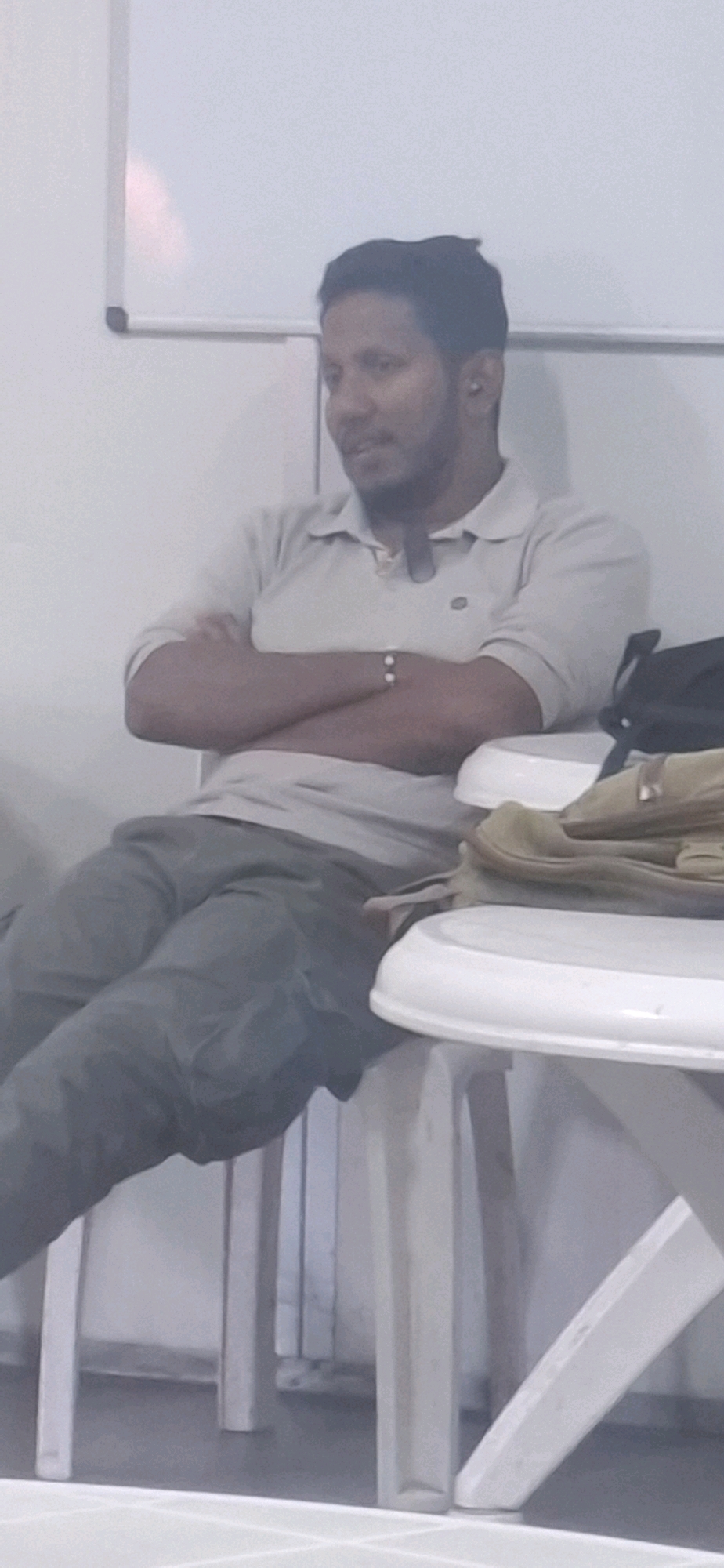 A man is sitting on a white plastic chair in front of a white table. He is wearing a light-colored polo shirt and grey pants. His arms are crossed and he is looking to his right. There is a black bag on the table to his right, and a light brown bag is also visible on the table. In the background, there is a white board on the wall and some other white chairs. The overall scene appears to be indoors, possibly in a classroom or meeting room, and the lighting is somewhat dim. The image does not provide enough information to determine the specific location within Kiryat Gat, Israel.