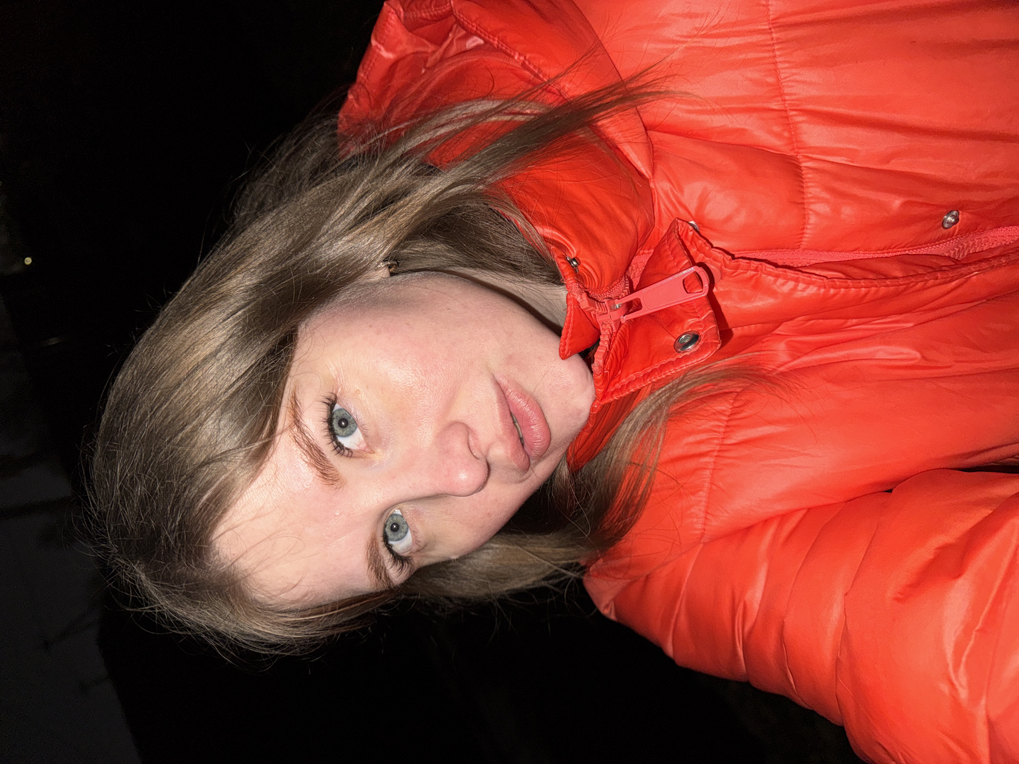 A young woman with fair skin, light brown hair, and blue eyes is wearing a bright orange puffer jacket. The jacket is zipped up to her neck. The background is predominantly black, suggesting a nighttime or low-light environment. The image appears to be taken at an angle, with the woman's face tilted towards the camera. The lighting is uneven, highlighting certain parts of her face and jacket while leaving other areas in shadow. The location is Горный, Russia.