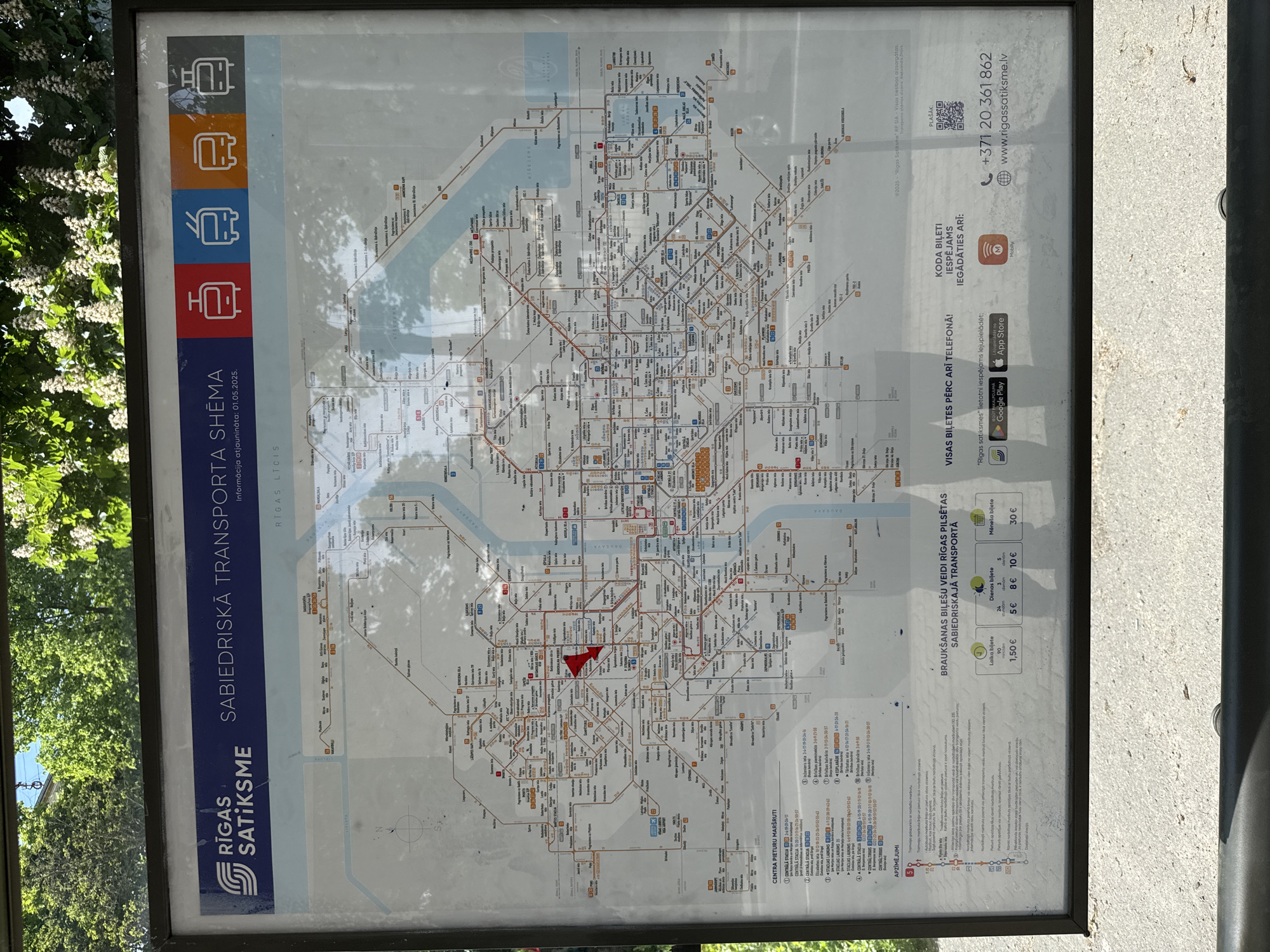 The image shows a public transport map of Riga, Latvia. The map displays the routes of buses, trams, and trolleybuses throughout the city. The map is mounted on a sign, and various ticket options and related information are provided, including QR codes to purchase tickets and apps to use for ticket purchase.