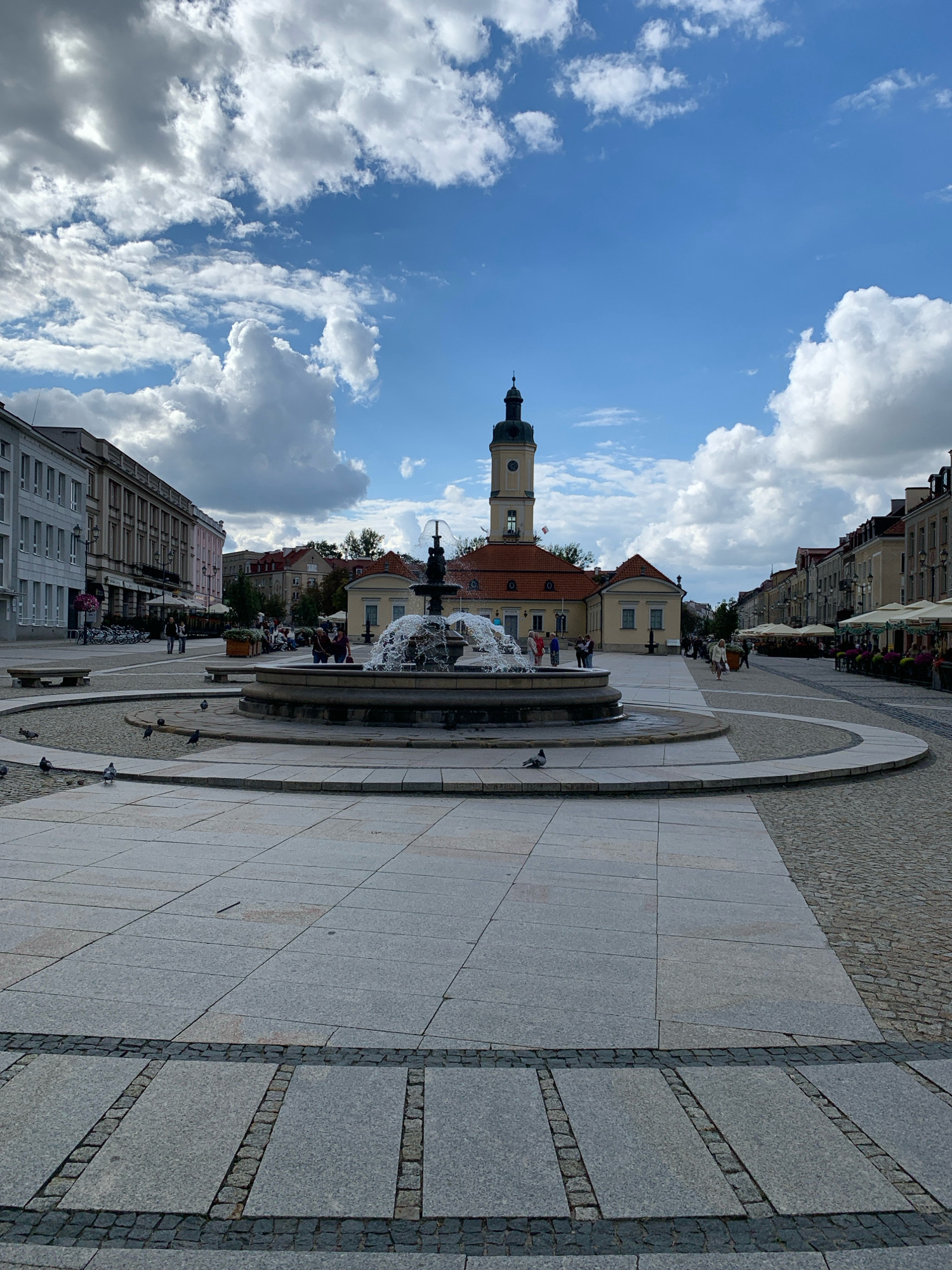 The image depicts a public square in Białystok, Poland, featuring a large fountain at its center. The square is paved with large, light gray stone slabs, bordered by cobblestones. Surrounding the fountain are several pigeons walking on the pavement. In the background, a distinct building with a clock tower, painted in yellow with a red roof, stands prominently. To the left of the tower, a modern, light-colored building with many windows lines the square. To the right, a row of traditional buildings with red roofs and awnings extends along the street. Several people are visible in the distance, some seated near the fountain and others walking along the street lined with outdoor seating for cafes. The sky is blue with scattered white clouds, indicating a partly cloudy day. The scene appears to be a lively, urban public space.