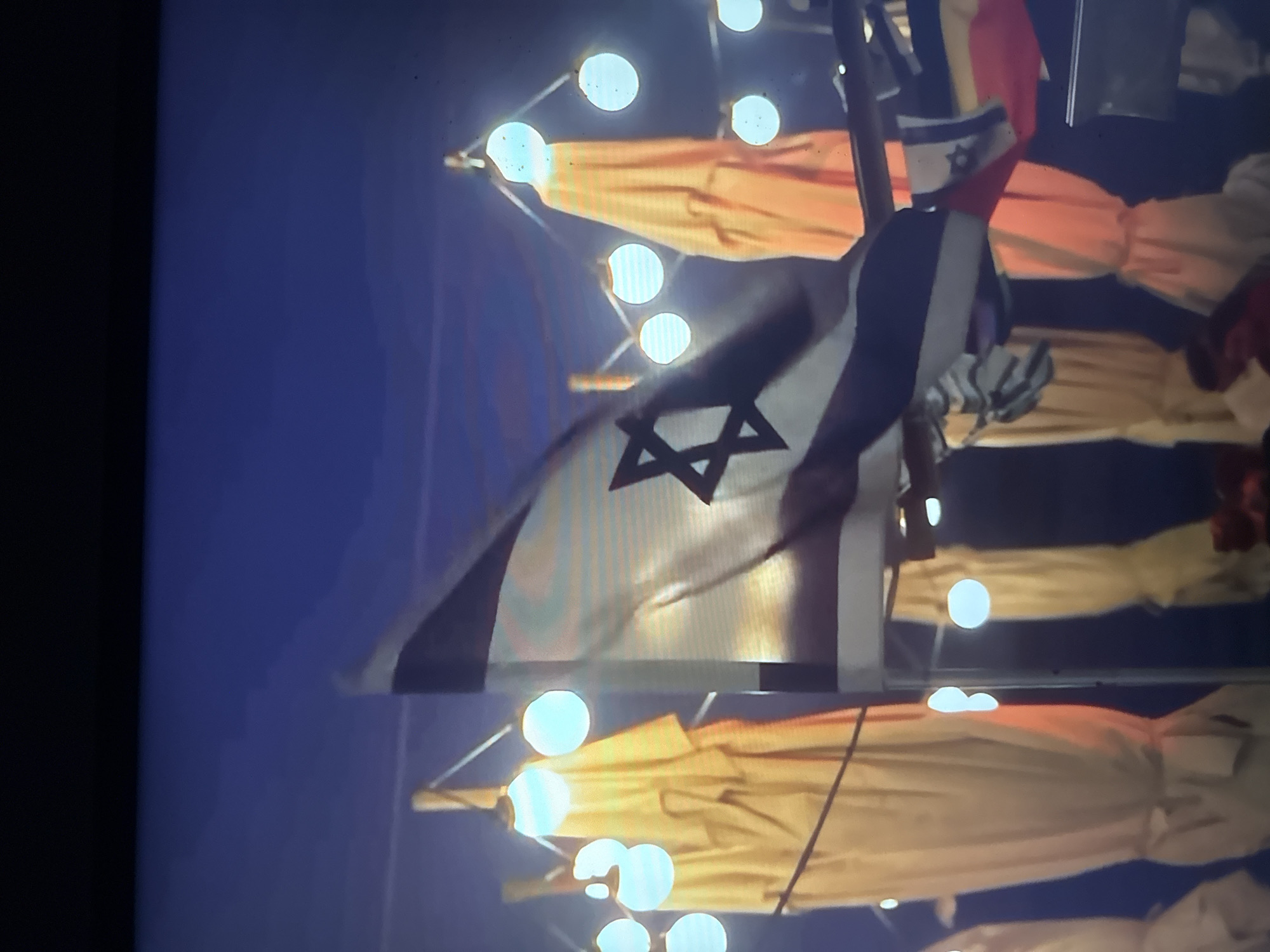 The image depicts an outdoor event at night, likely in Herzliya, Israel, given the context. The central focus is a flag of Israel, partially unfurled and illuminated by bright, circular lights. Behind the Israeli flag, other flags, some appearing to be of red and yellow colors, are visible, along with what look like folded tents or awnings, also illuminated. The lighting suggests the event is taking place after dark, with the bright lights creating a festive or ceremonial atmosphere. The overall scene is dark with bursts of light, emphasizing the flags and structures. There is no visible text in the image.