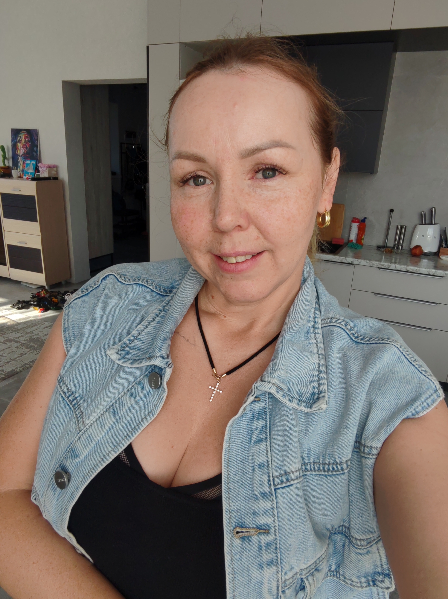 A light-skinned woman with reddish-blonde hair, likely in her late 30s to mid 40s, is taking a selfie in what appears to be a modern apartment or house interior. She is wearing a light blue denim vest over a black top, with a gold-colored cross pendant on a black cord necklace. She also sports gold hoop earrings. Her face is freckled.
Behind her, the background suggests a kitchen area with light gray cabinets and stainless-steel utensils. To the left is a hallway leading to another room where a bicycle is visible. There's also a dresser with what appears to be an art piece and a few decorative items on top.
Given the request to include the location "Chaikovsky, Russia," one can infer that the woman is located in Chaikovsky, Russia. There is no definitive information to confirm this is actually the location.
