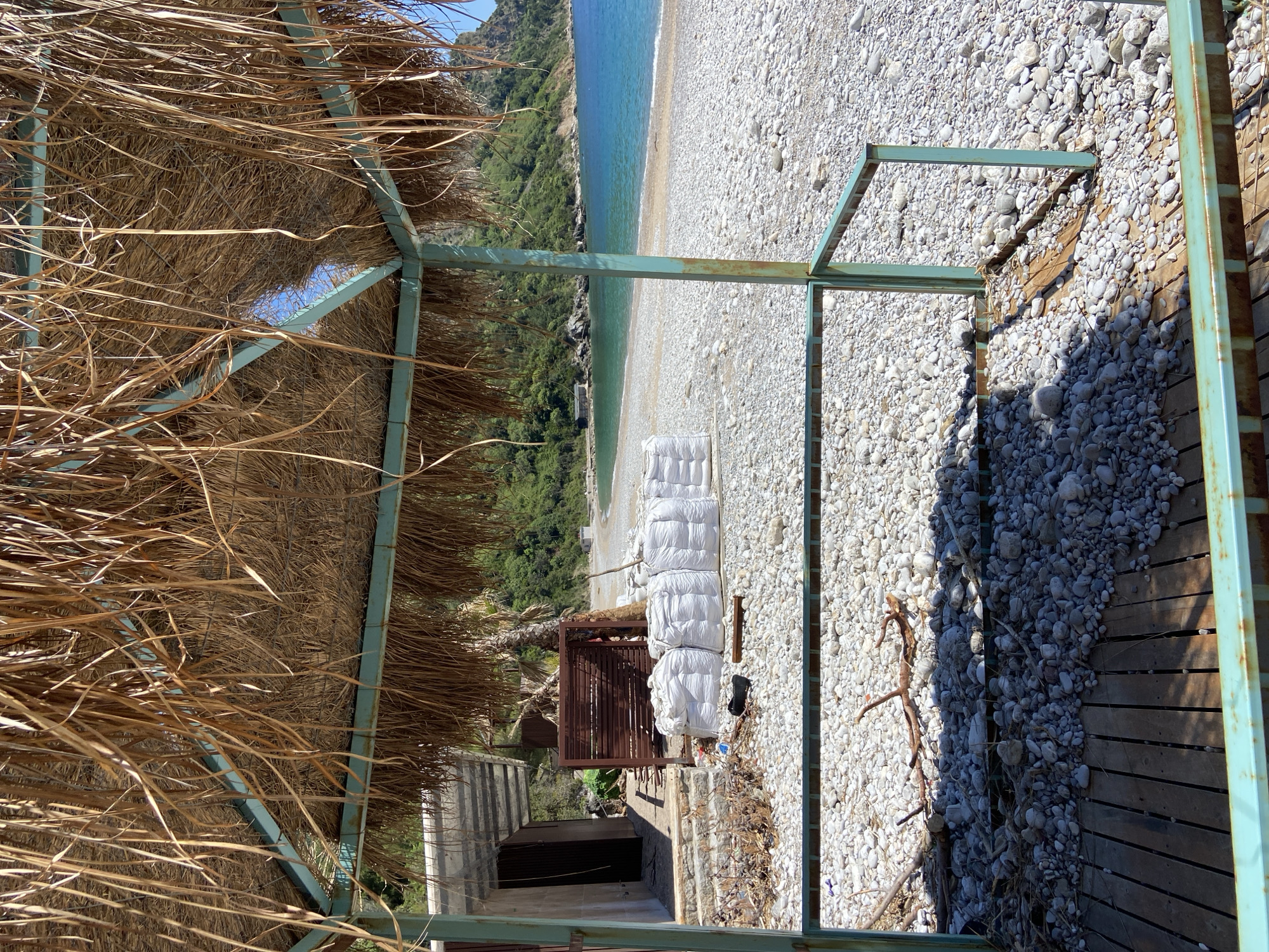 The image depicts a beach scene in Prijevor, Montenegro, viewed from under a straw-roofed structure with a blue-green metal frame. The beach is covered in smooth, white pebbles, sloping down towards a turquoise sea. In the middle distance, there are several white, fabric-covered items (possibly stacked cushions or mattresses) situated near a brown, slatted fence-like structure. A dark, open doorway is visible beneath a concrete structure behind the fence. Vegetation, likely trees and shrubs, covers the hillside in the background. A wooden deck sits next to the pebbles of the beach.