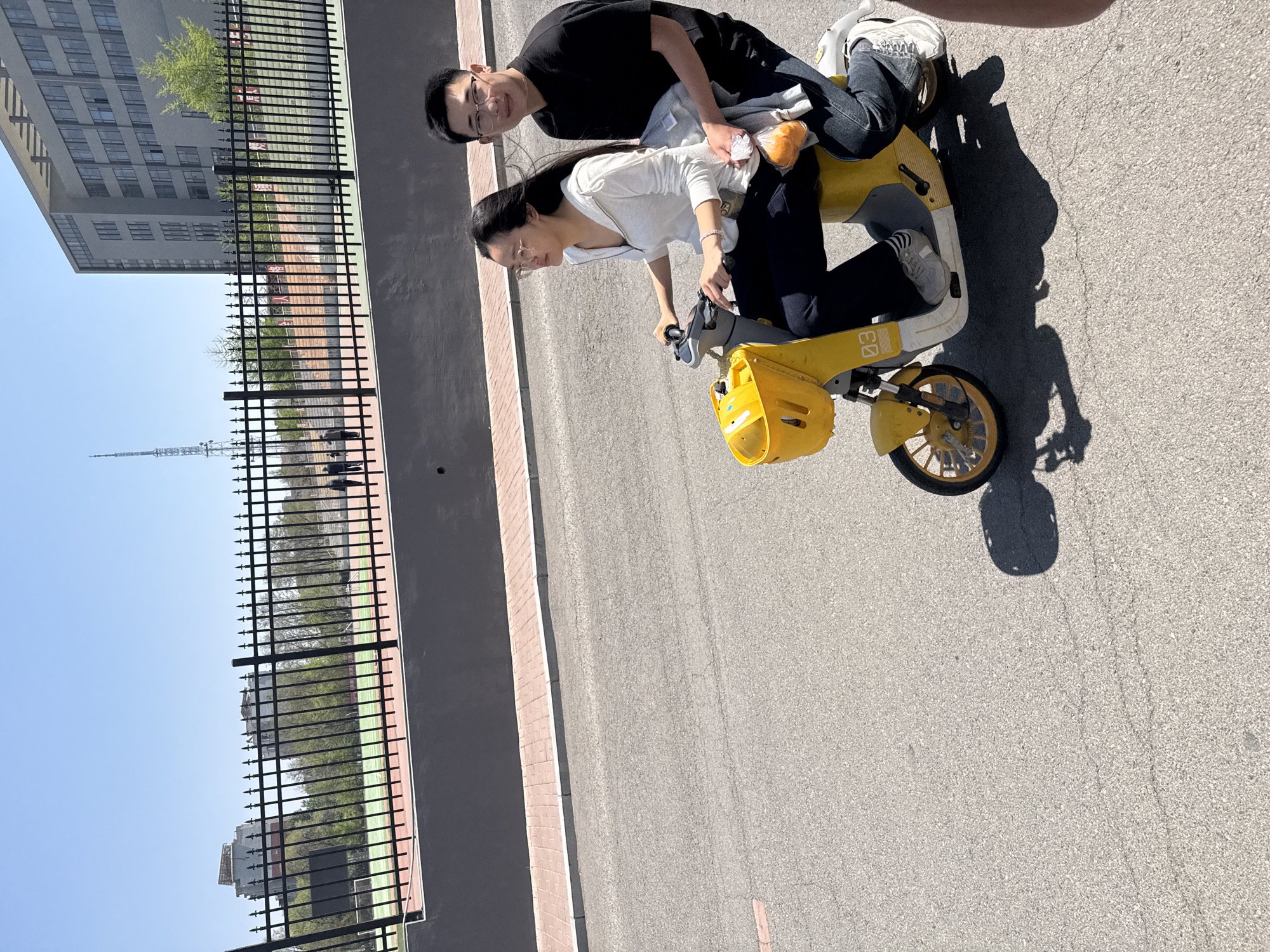 A brightly lit, daytime image shows two people on a yellow scooter-like vehicle in Zhongtun, China. A person with dark hair and glasses, wearing a white shirt and black pants, is operating the vehicle, while another person with short dark hair and glasses, wearing a black shirt and jeans, sits behind as a passenger. The vehicle has a yellow basket in the front. They are on a paved road with a sidewalk and a black metal fence running alongside it. Beyond the fence, there is a green space with buildings and a tall communications tower visible in the background. The sky is clear and blue.