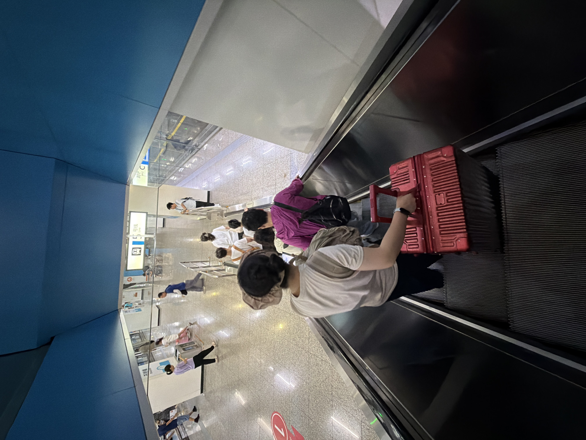 In the Jiulongpo District of China, this image captures a scene on an escalator within a subway station. The escalator is moving downwards, and people are descending on it. The foreground shows a person from behind, wearing a light-colored t-shirt and black pants, holding the handle of a red, hard-shell suitcase. Another person in a purple jacket with a black backpack is just ahead of them on the escalator. In the background, the subway platform is visible with several other individuals, some walking and others standing. The station features a bright, modern design with blue and white panels on the ceiling and walls. Signage in Chinese characters is present, indicating station information. The floor is tiled and reflects the overhead lighting. A red circular marker with a white "1" is visible on the floor in the mid-ground.