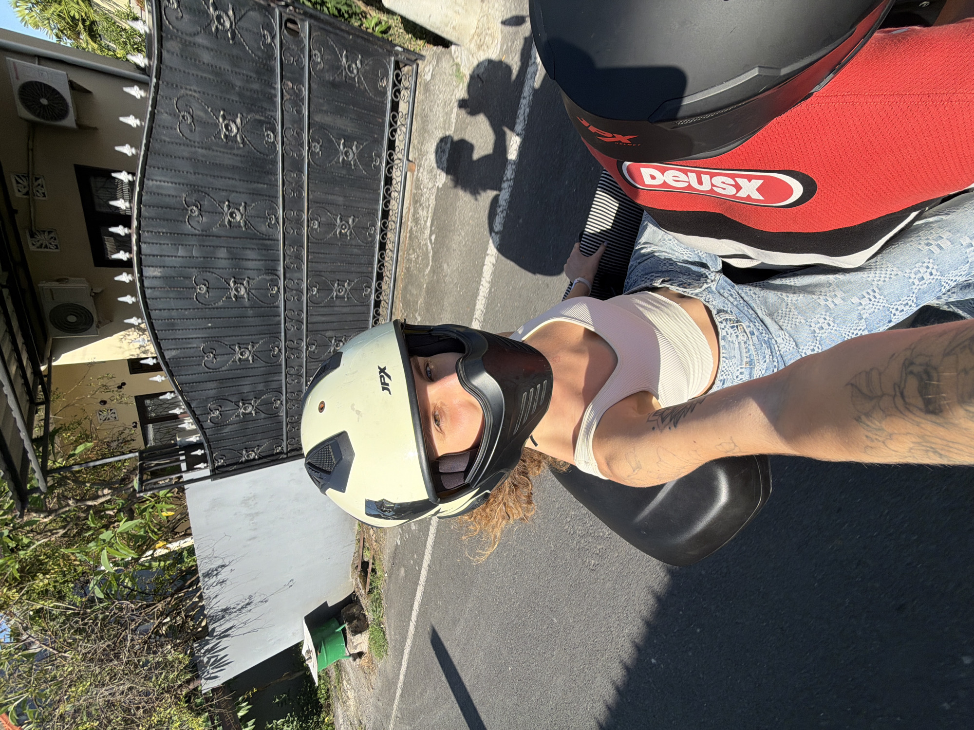 A person is riding a motorcycle down a paved street in North Kuta, Indonesia, on a sunny day. They are wearing a cream-colored helmet with black accents and a black face shield. The person is also wearing a white tank top and distressed denim jeans. Their left arm, visible in the foreground, is tattooed with various designs. The motorcycle has a black seat and a red and black cover with the "Deus Ex" logo on the rear passenger seat. In the background, there is a tall black metal gate with ornate patterns, a light-colored wall, trees with green foliage, and a building with two air conditioning units. The street has white lane markings and a shadow of the rider and motorcycle is cast on the pavement.