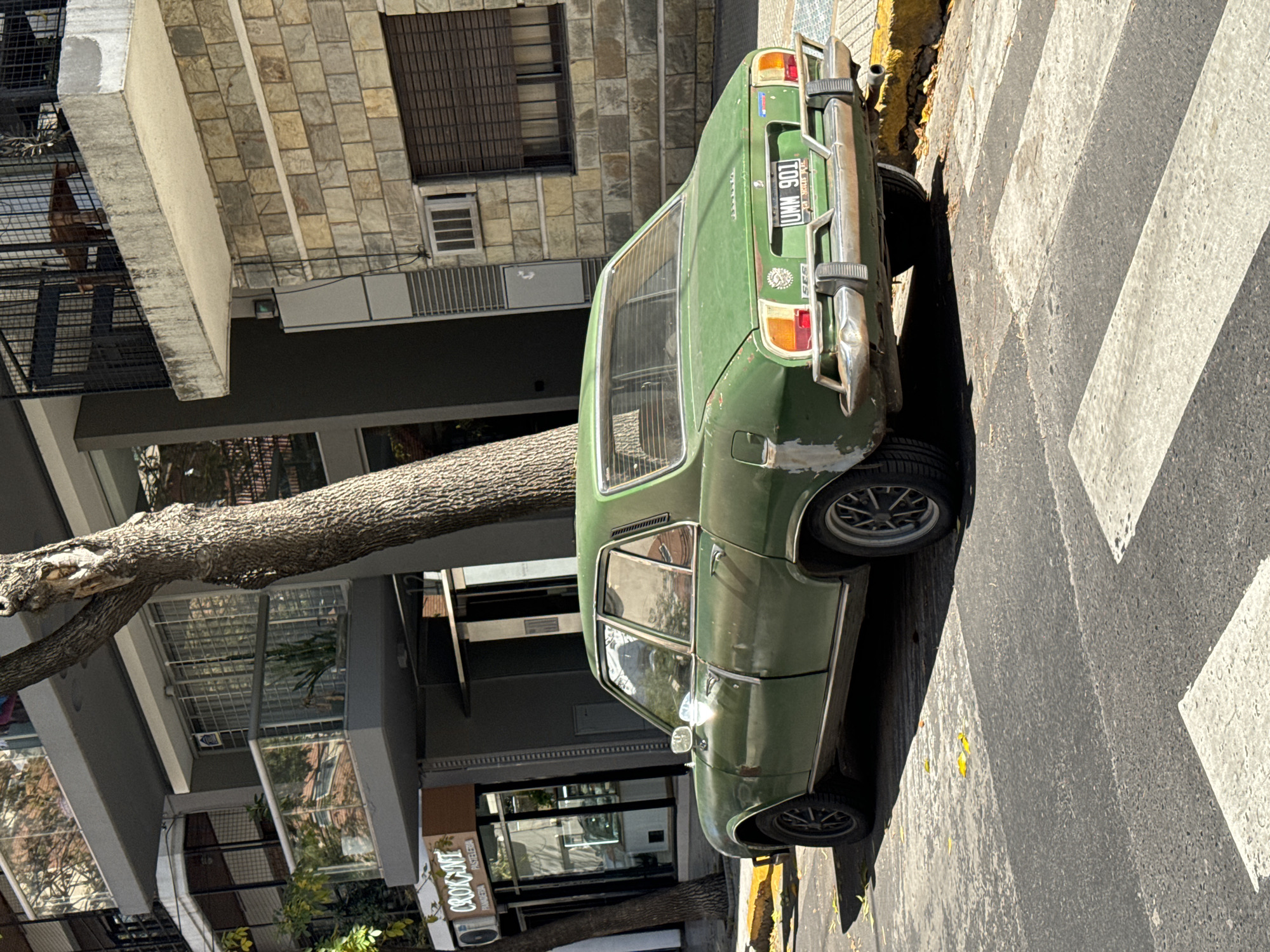 A medium shot depicts an olive green 1970s IKA Renault Torino parked on a crosswalk near a tree and building on a city street. The car is angled to the right with the rear facing the camera, the license plate reading "UNM 901." The car is parked on the edge of the sidewalk, which is lined with a yellow curb. A tree trunk obscures part of the car and building behind. The building has a sign reading "CROQUANT PASTELERIA PANADERIA." The crosswalk is painted with alternating white and gray stripes. The building is constructed of beige stone blocks. The scene is brightly lit, suggesting daytime. Based on the license plate and architectural style, the location is likely Buenos Aires, Argentina. There are no visible people.