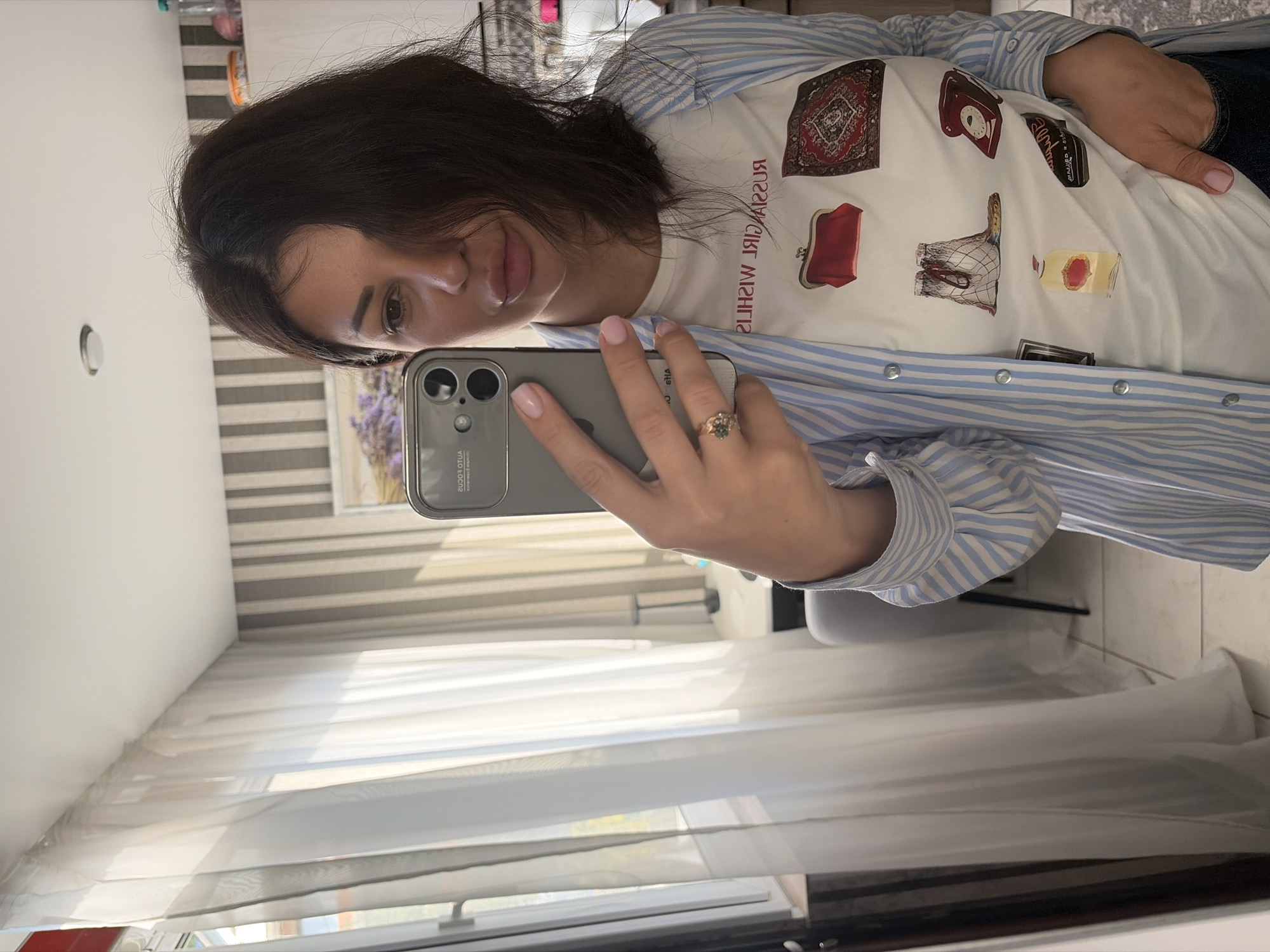 A young woman in Sochi, Russia, captures a mirror selfie indoors during the day, indicated by the natural light. She holds a modern smartphone, likely an iPhone, in her right hand, which features light pink nail polish and a gold-toned ring with a green stone. Her dark hair frames her face, and she looks directly into the camera with a gentle expression.
She is dressed in a white t-shirt adorned with various printed images: a red and black patterned rug, a red vintage purse, a black rotary telephone, a perfume bottle with a red top, and a small piece of folded clothing. The t-shirt also displays the reversed text "RUSSIAN GIRL WISHES." Over this, she wears an open, light blue and white striped button-down shirt with silver snap buttons. Dark pants or jeans are partially visible on her lower body.
The indoor setting features a background with a striped wall or blinds and a large window covered by sheer white curtains, allowing sunlight to diffuse into the room. A subtle reflection in her phone's screen shows more of the room, including what might be a plant or flowers near the window.