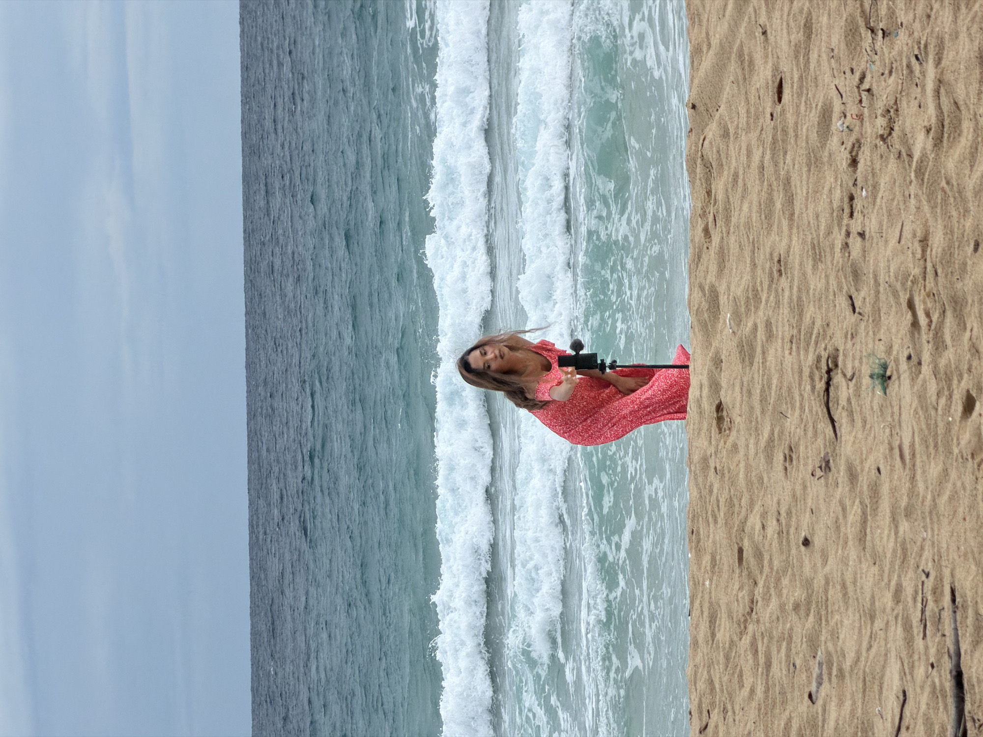 A young woman in a red dress with white floral patterns is crouched on a sandy beach, reaching for a black tripod with a device attached to it. She is facing away from the viewer, towards the ocean. The ocean water is a turquoise color with white waves crashing on the shore. The sky is overcast and a light gray color. The beach is covered in light brown sand with some scattered debris. The image is taken at an eye-level perspective, with the woman positioned in the lower half of the frame. The background shows the vast expanse of the ocean and the sky.