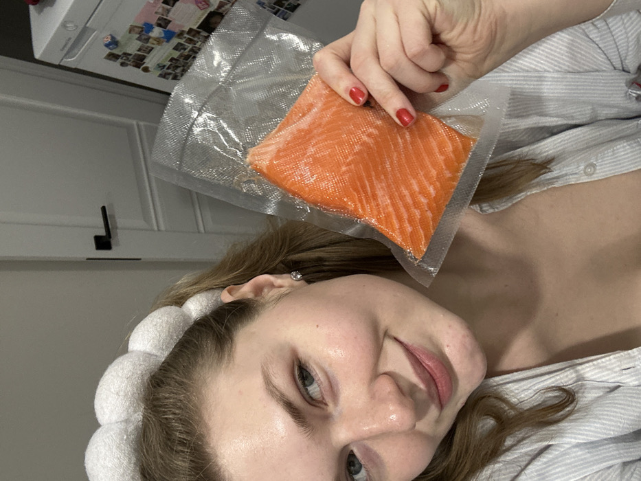 A woman with fair skin, light eyes, and a white, puffy headband is holding a package of salmon. The salmon is orange and sealed in clear plastic. The woman has red nail polish on her fingernails. She's wearing a light-colored, striped shirt. In the background, there is a white refrigerator with pictures on it and a white cabinet. The overall lighting is soft and diffused.