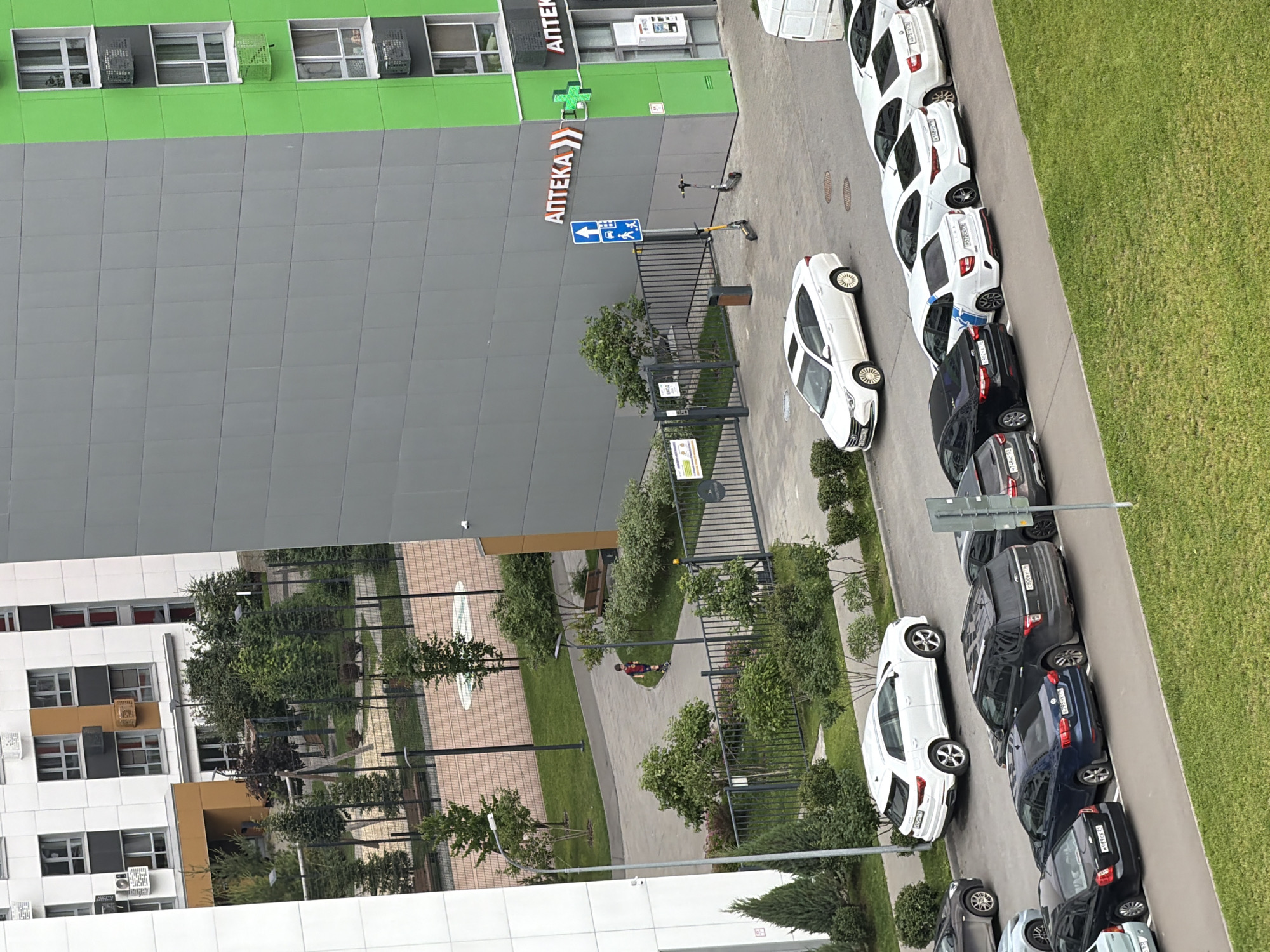 An aerial view shows a parking lot filled with cars parked on a paved surface next to a green lawn. The cars are of various colors, including white, black, and blue. In the background, there is a modern building with a gray and green facade. A sign with the word "АПТЕКА" (Pharmacy) in large red letters is visible on the building, along with a green cross symbol. Two electric scooters are parked on the sidewalk near the building. Further in the background, another building with white and orange panels is visible, with a courtyard featuring a playground and trees. A person on a scooter is seen in the courtyard. The scene appears to be in a residential or commercial area.