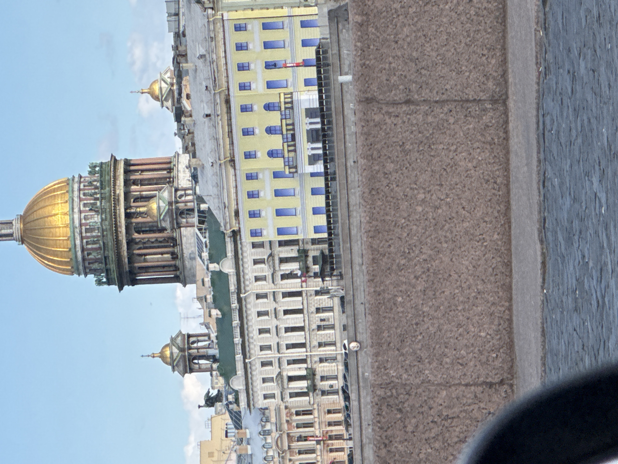 The image shows a view of St. Isaac's Cathedral in Saint Petersburg, Russia, along with adjacent buildings. The cathedral's golden dome is prominently featured against a blue sky. A portion of a bridge or embankment is visible in the foreground, suggesting the photo was taken from that vantage point. The surrounding buildings display varied architectural styles and colors. No people are directly observable in the scene.